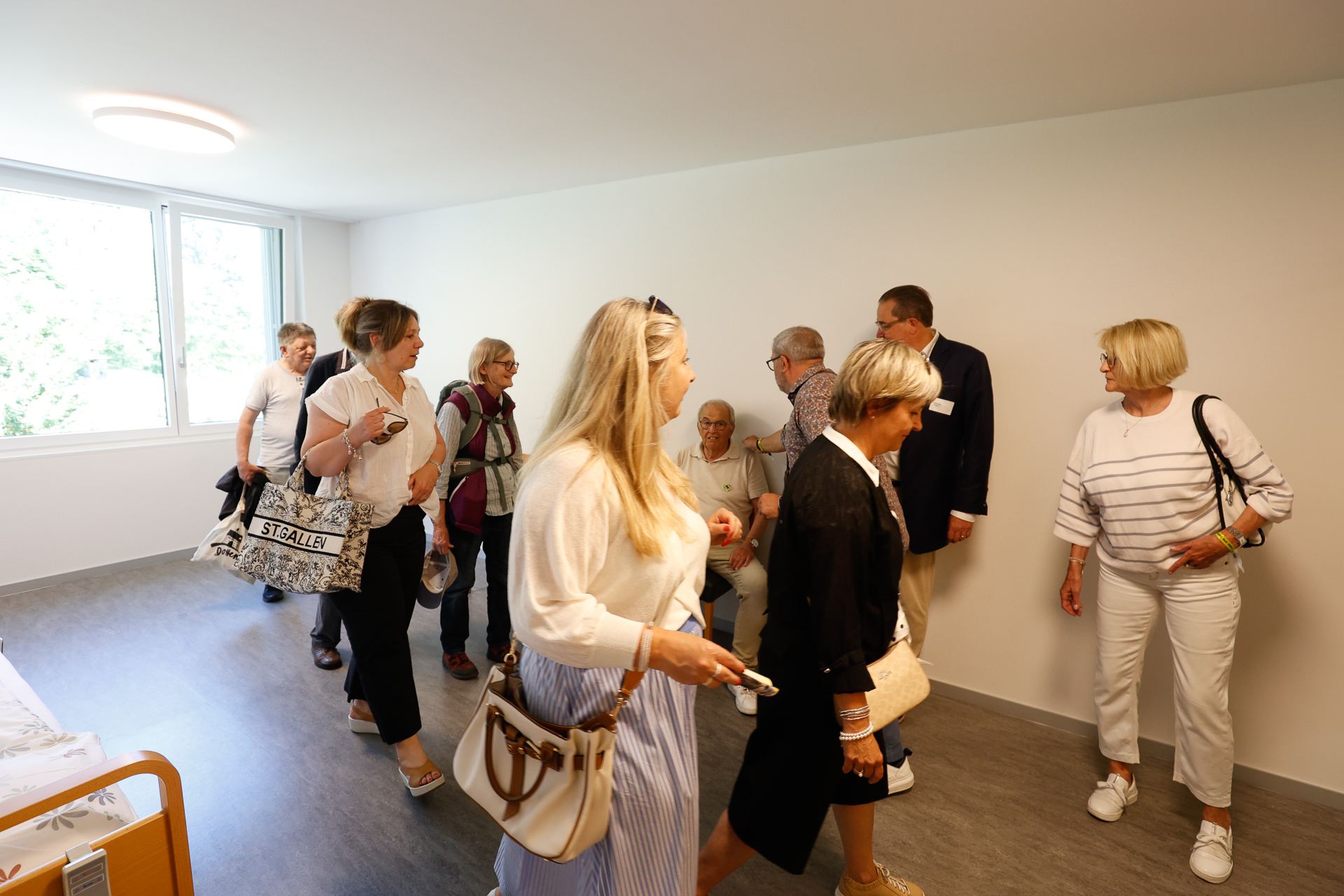 Eine Gruppe von Erwachsenen, sowohl Männer als auch Frauen, geht in einem hellen, spärlich möblierten Raum mit einem großen Fenster umher und unterhält sich. Einige stehen, während andere auf Stühlen an der Wand sitzen.