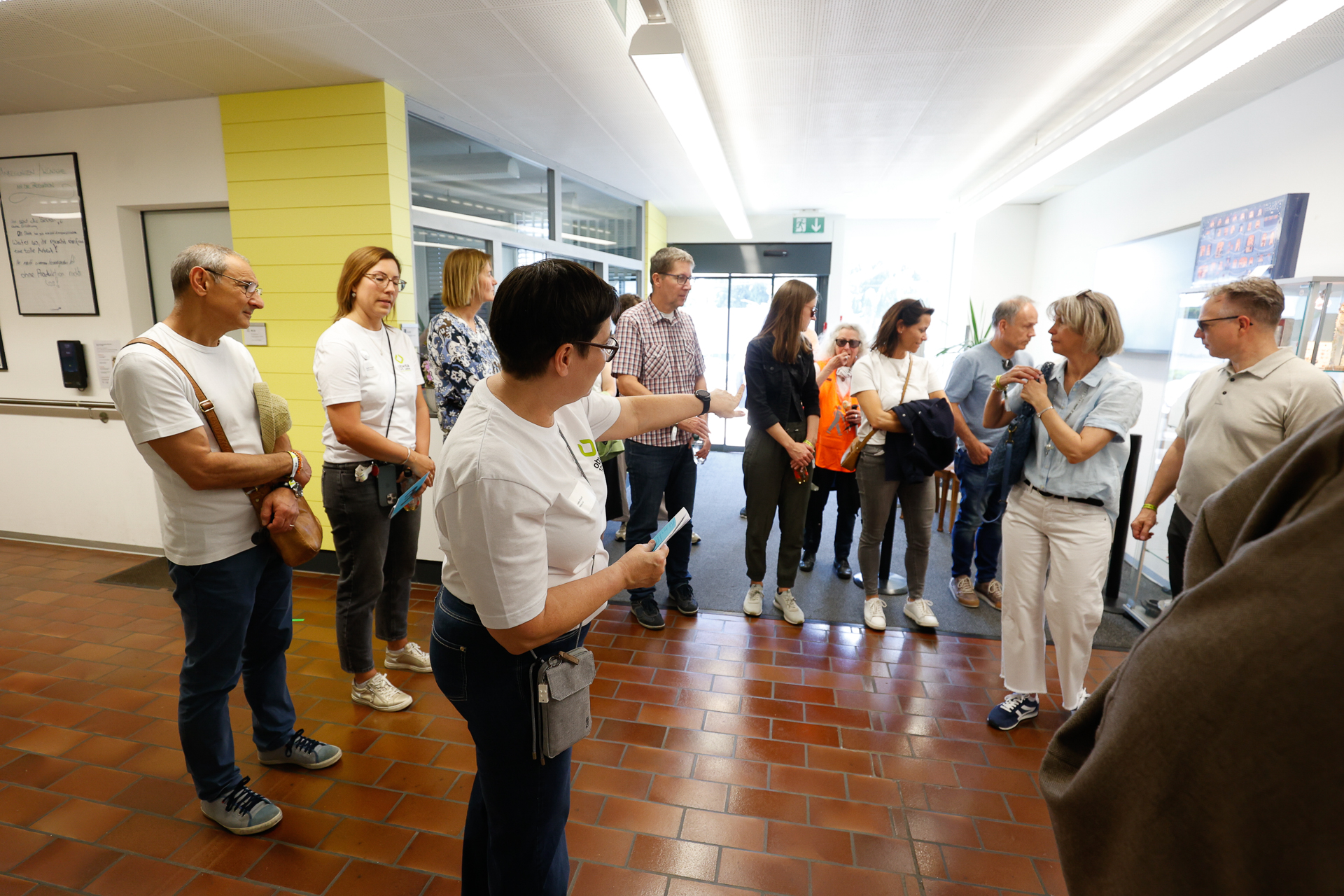 Eine Gruppe von Erwachsenen steht in einem hellen Flur mit Fliesenboden. Eine Frau im Vordergrund gestikuliert, während sie zu der Gruppe spricht, die leger gekleidet ist und zuzuhören oder sich zu unterhalten scheint.