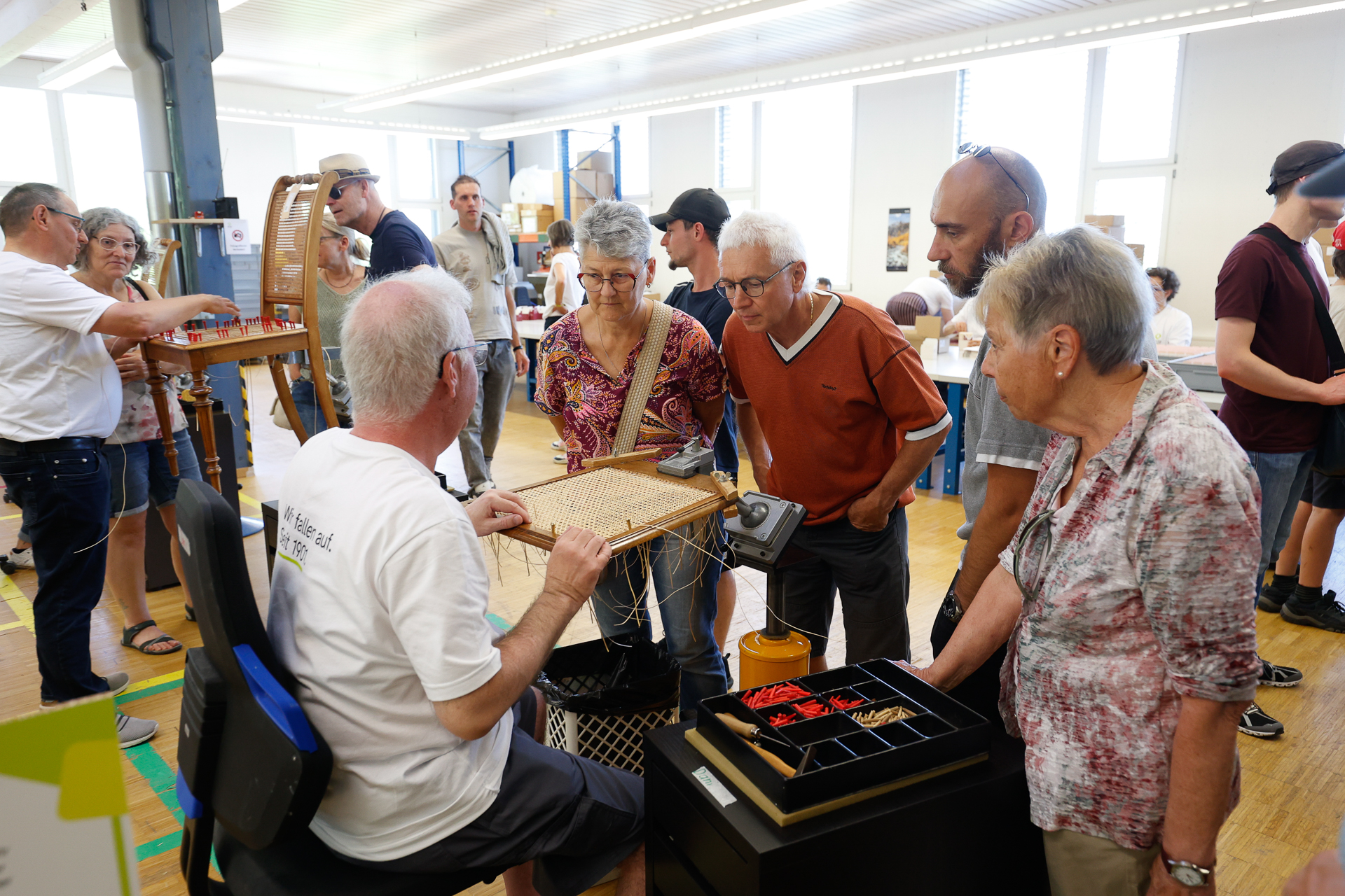 Eine Gruppe von Menschen versammelt sich um einen Mann, der bei einem Workshop oder einer Vorführung einen Sitz flechtet; andere schauen zu und unterhalten sich in einem hellen Innenraum mit großen Fenstern.
