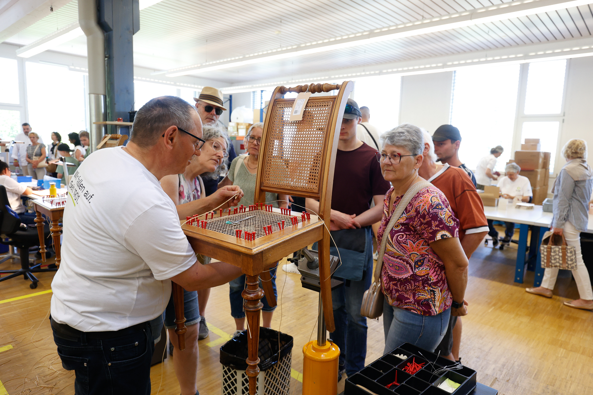 Eine Gruppe von Menschen versammelt sich um einen Mann, der in einem hellen, belebten Innenraum – möglicherweise einem Workshop oder Ausstellungsraum – ein Brettspiel oder ein Gerät mit roten und weißen Spielsteinen vorführt.