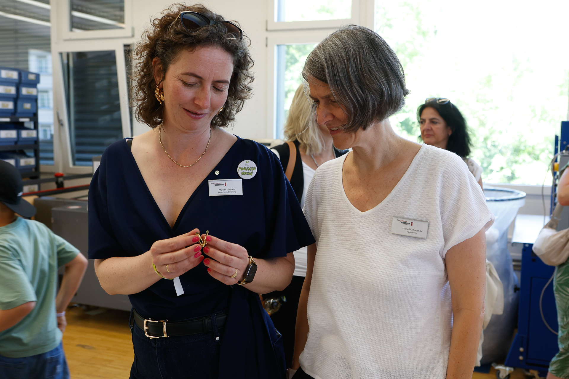 Zwei Frauen mit Namensschildern stehen in einem Raum; die eine hält einen kleinen Gegenstand in der Hand und lächelt, während die andere sie dabei beobachtet. Im Hintergrund sind weitere Personen in einem hellen Raum mit großen Fenstern zu sehen.