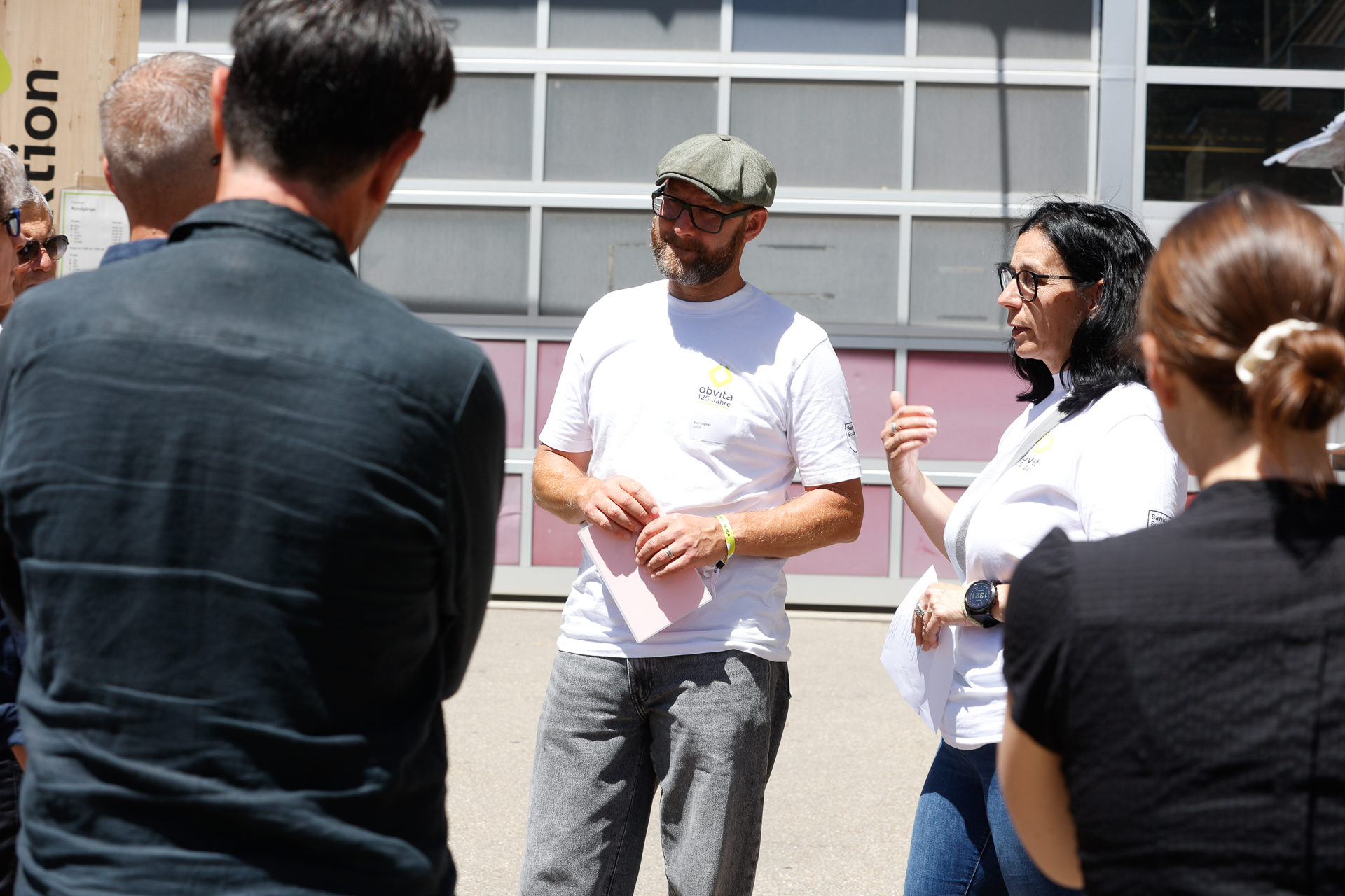 Ein Mann und eine Frau in weißen T-Shirts stehen draußen und sprechen mit einer kleinen Gruppe von Menschen. Der Mann hält einige Blätter in der Hand, und die Gruppe hört aufmerksam zu. Im Hintergrund ist ein Gebäude mit großen Fenstern zu sehen.