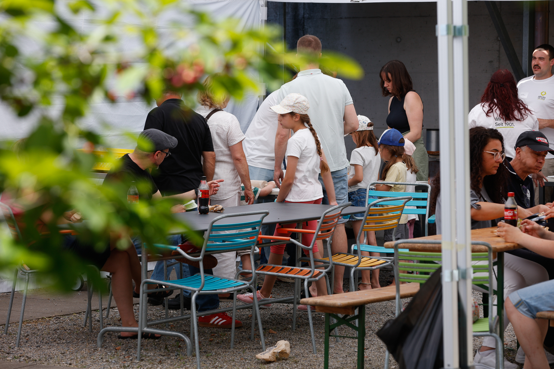Eine Gruppe von Menschen, darunter Kinder und Erwachsene, sitzt und steht im Freien um bunte Tische und Stühle herum. Einige trinken Getränke aus Flaschen. Grüne Blätter rahmen die Szene teilweise ein. Es scheint sich um ein ungezwungenes geselliges Beisammensein zu handeln.