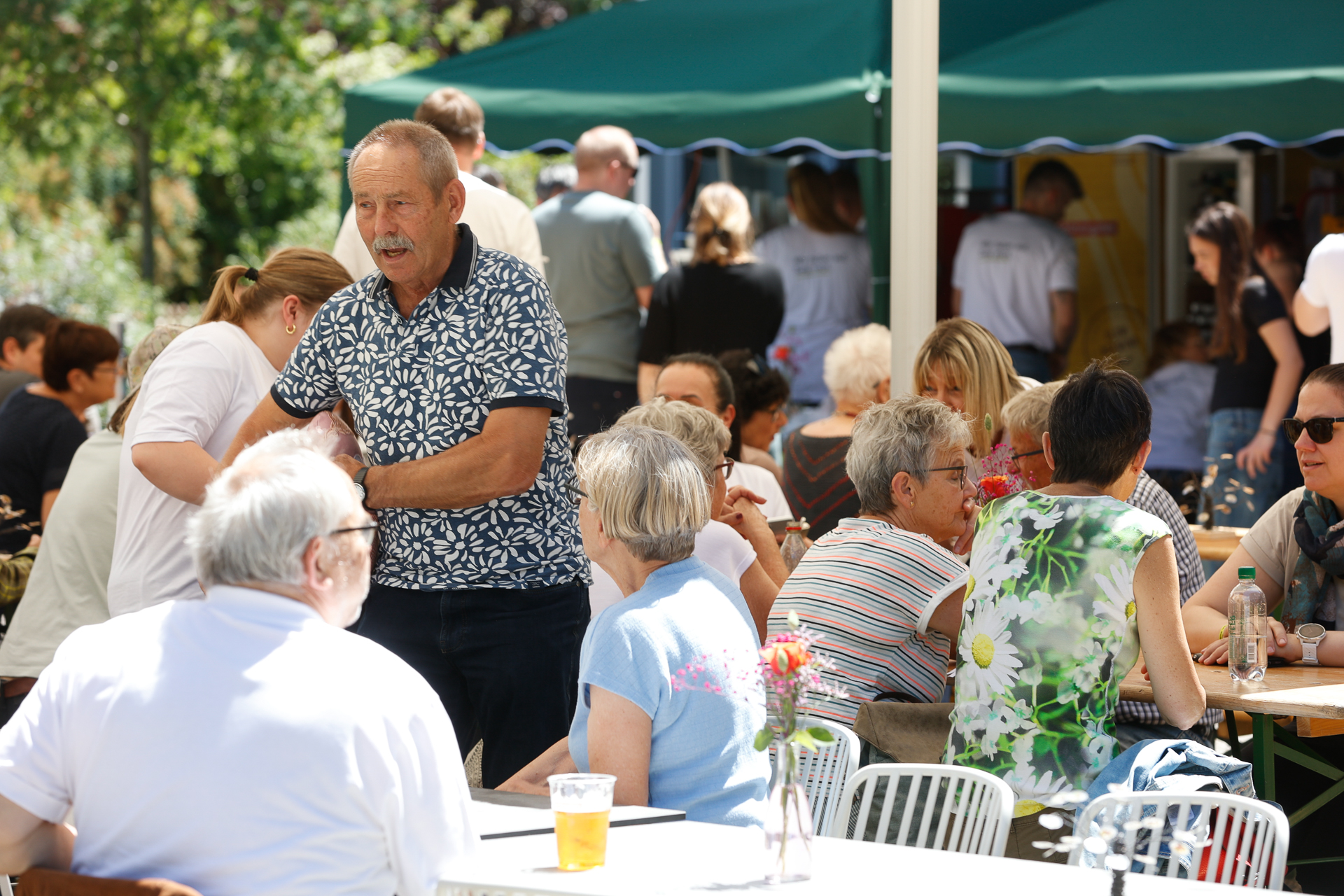 Menschen unterschiedlichen Alters sitzen an Tischen im Freien unter grünen Markisen, unterhalten sich und genießen an einem sonnigen Tag ihre Getränke. Einige stehen und unterhalten sich, während andere sitzen. Blumen schmücken die Tische.