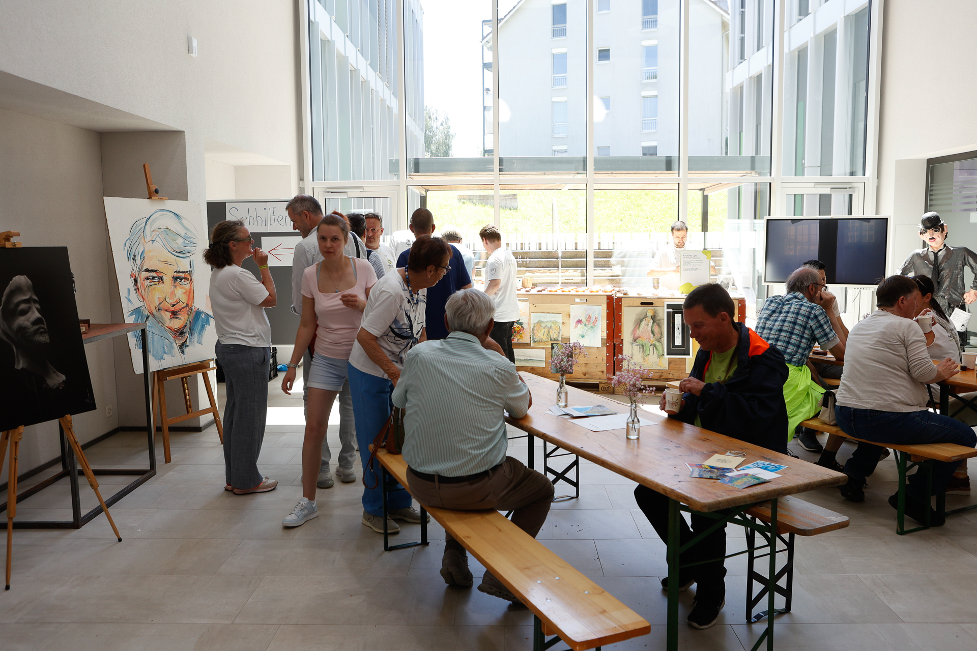 In einem hellen, modernen Raum mit großen Fenstern versammeln sich Menschen zu einer Kunstveranstaltung. Einige sitzen an Holztischen, während andere neben Staffeleien stehen, auf denen Porträts und Gemälde ausgestellt sind. Auf den Tischen liegen Malutensilien und Broschüren.