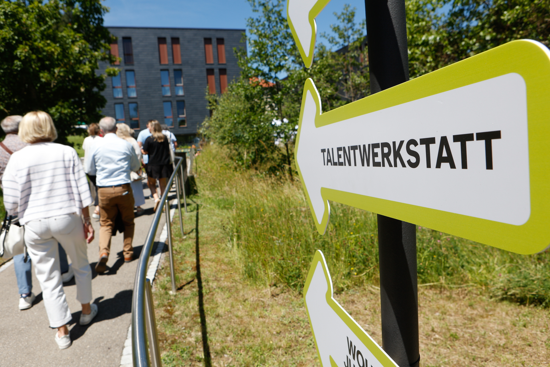 Eine Gruppe von Menschen geht im Freien einen Weg entlang in Richtung eines modernen Gebäudes und kommt dabei an einem Schild mit Pfeilen vorbei, auf dem „Talentswerkstatt“ steht. An diesem sonnigen Tag säumen grünes Gras und Bäume den Weg.