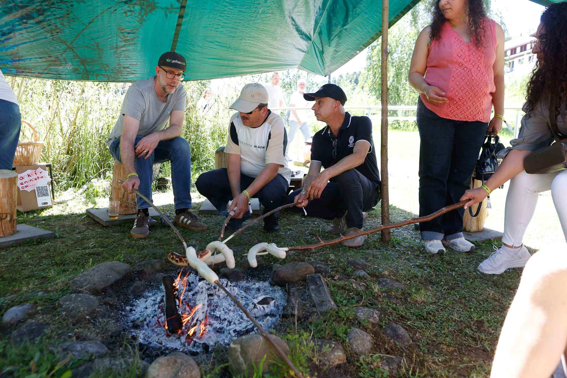 Menschen sitzen unter einer grünen Plane um ein Lagerfeuer herum und rösten Brotteig auf Stöcken über den Flammen. Die Gruppe wirkt begeistert und genießt die Aktivität im Freien.