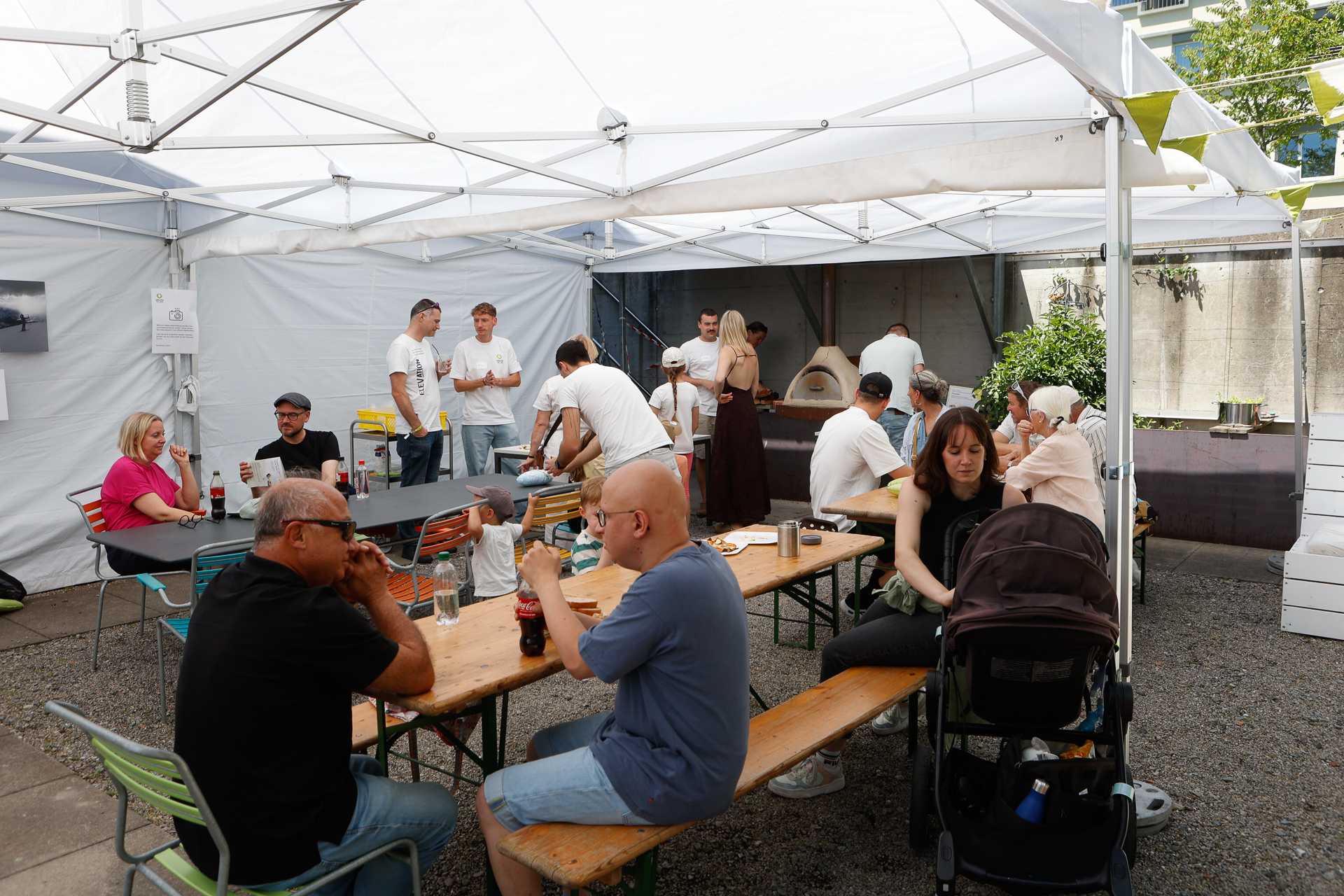 Die Menschen versammeln sich unter einem weißen Zelt, sitzen an Picknicktischen und unterhalten sich. Einige essen, während andere in der Nähe eines Pizzaofens im Freien Essen zubereiten. Eine Frau sitzt mit einem Kinderwagen da; die Atmosphäre ist ungezwungen und gesellig.