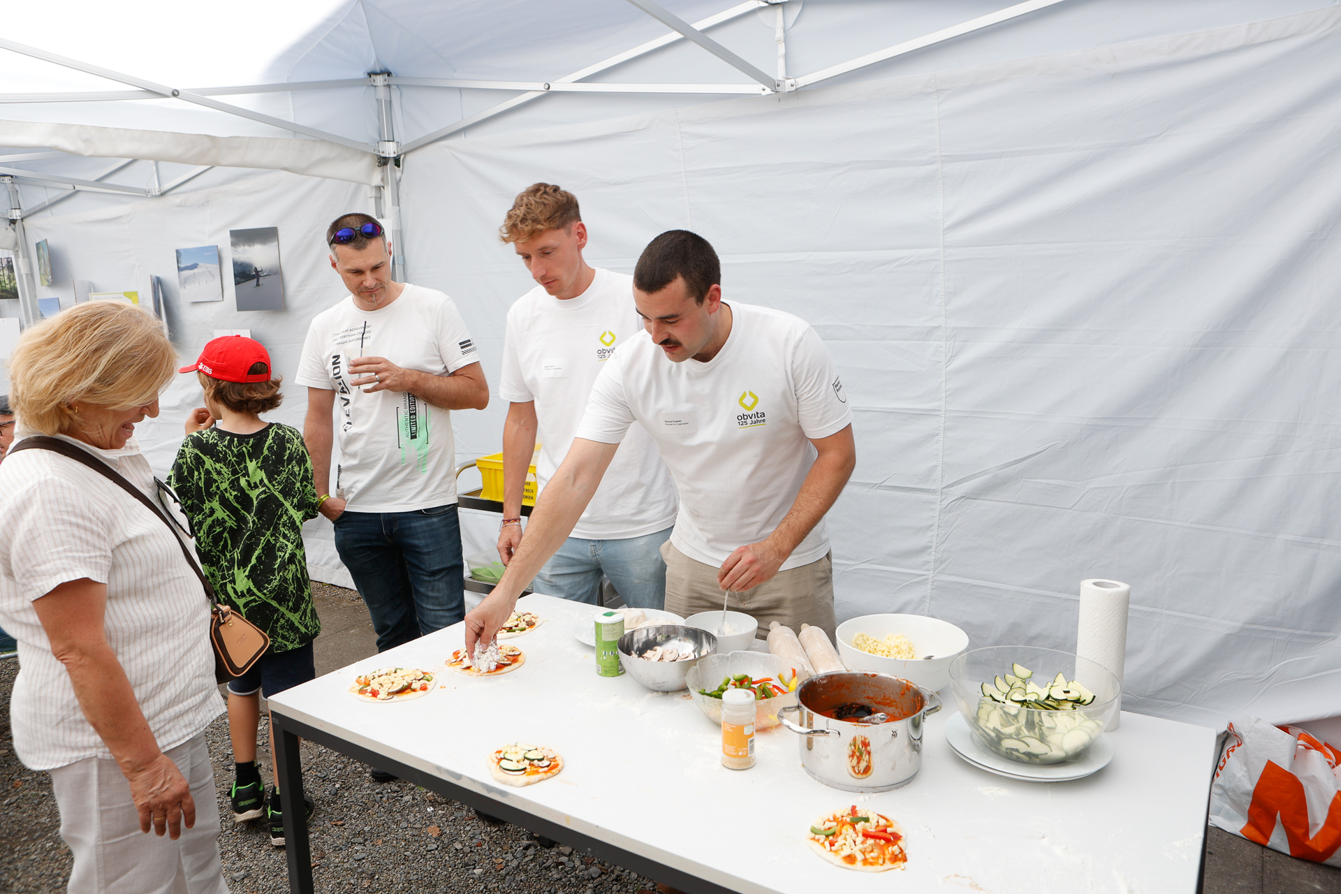 Vier Männer in weißen Hemden bereiten an einem Tisch unter einem weißen Zelt Mini-Pizzen zu, während eine Frau und ein Kind zuschauen. Auf dem Tisch stehen Pizzazutaten wie Soße, Käse und Gemüse. An der Zeltwand sind Fotos zu sehen.