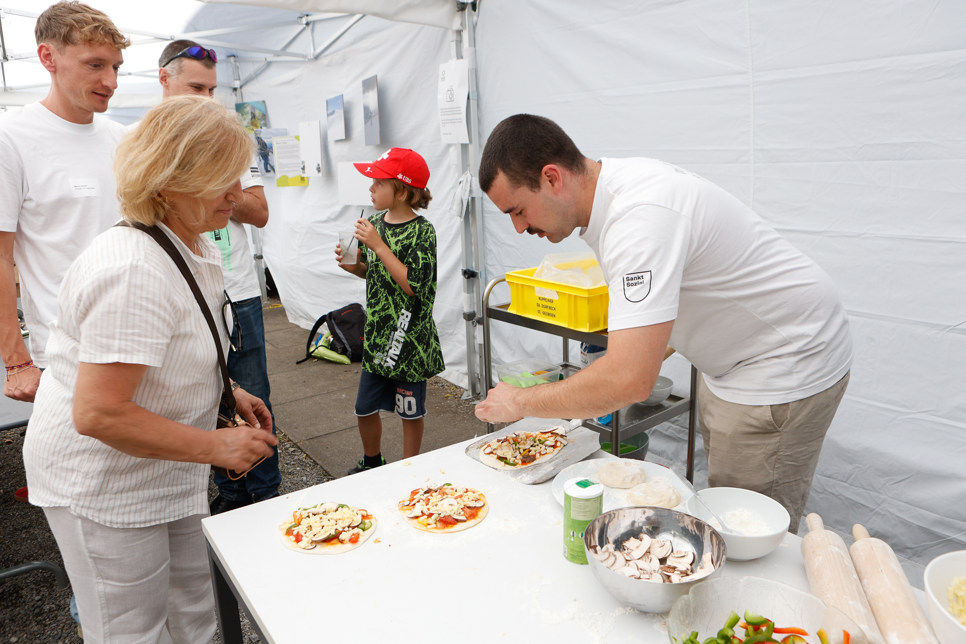 Ein Mann bereitet auf einem mit Zutaten bedeckten Tisch Pizzen zu, während eine Frau ihm dabei zusieht. Daneben stehen ein Junge mit einer roten Mütze und weitere Personen in einem weißen Zelt. Auf dem Tisch sind Pizzabeläge und Küchenutensilien zu sehen.