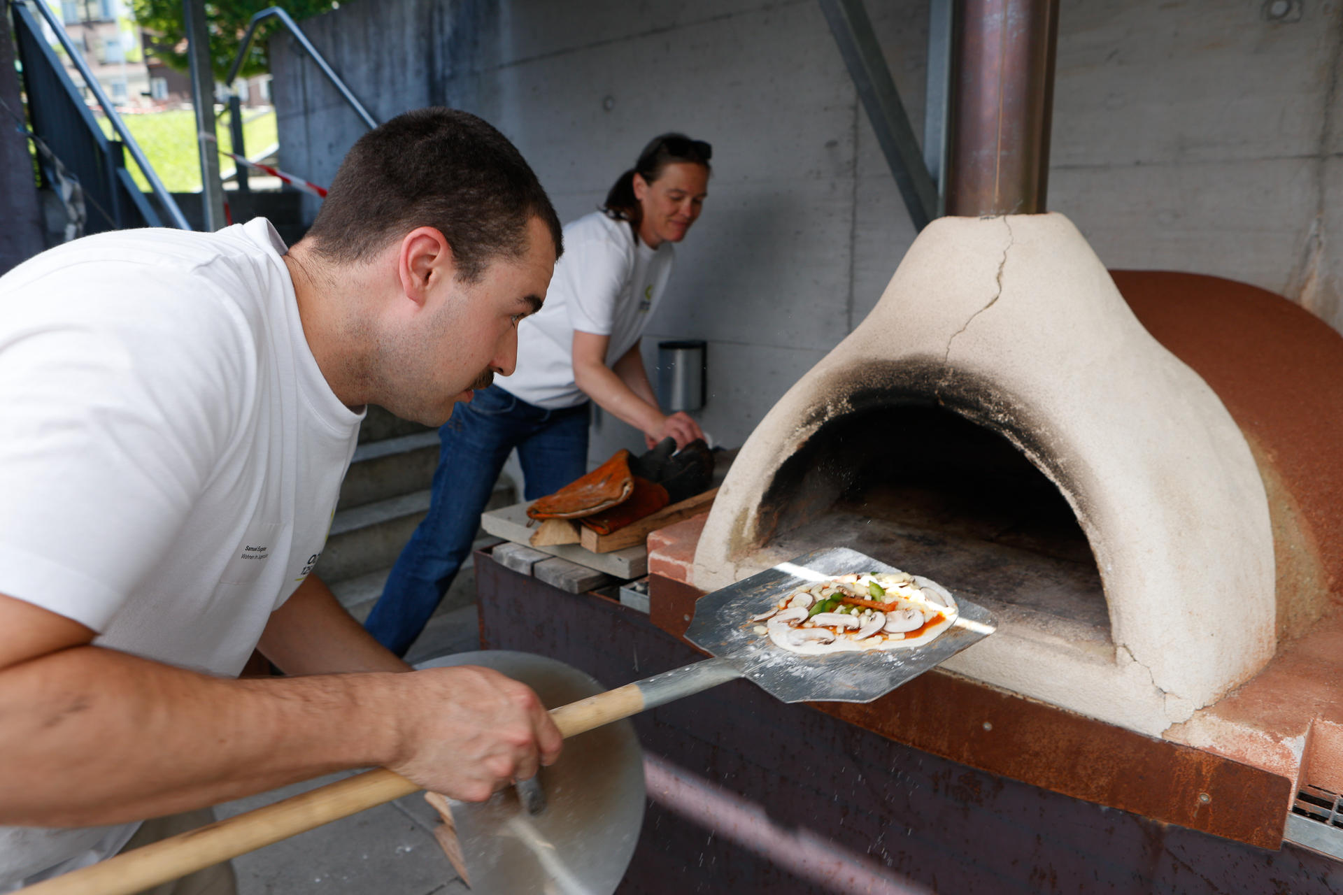Ein Mann schiebt mit einer Pizzaschaufel eine Pizza in einen Holzofen, während im Hintergrund eine Frau Essen zubereitet. Beide tragen weiße Hemden und arbeiten im Freien.