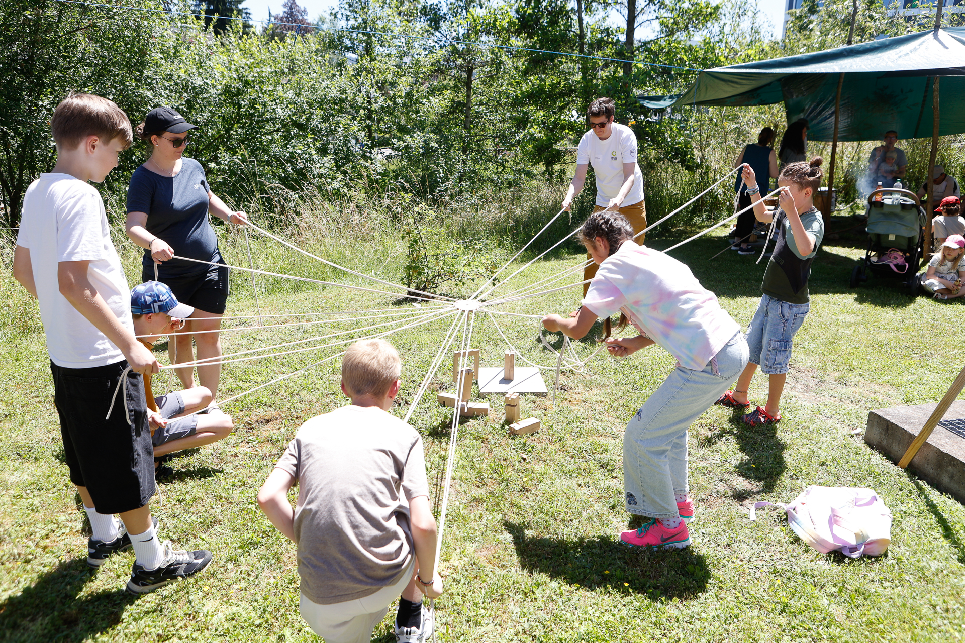 Eine Gruppe von Kindern und Erwachsenen arbeitet an einem sonnigen Tag im Freien gemeinsam daran, Holzklötze mithilfe von Schnüren über das Gras zu bewegen, umgeben von Bäumen und schattenspendenden Zelten.