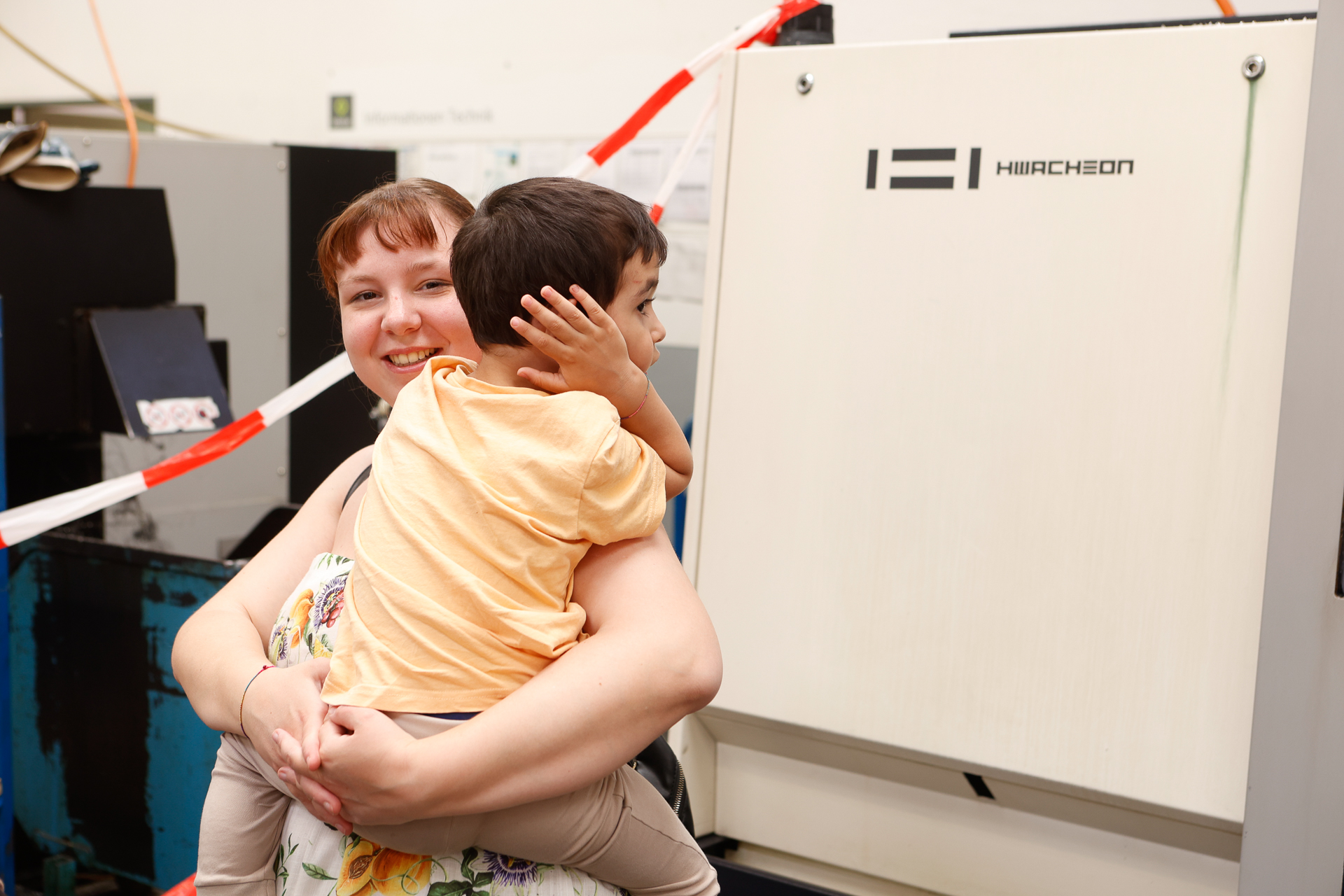 Eine lächelnde Frau hält ein kleines Kind im Arm, das sich die Hand vor ein Ohr hält, und steht neben einer weißen Industrieanlage mit der Aufschrift „HIIRCHON“. Im Hintergrund ist rotes und weißes Absperrband zu sehen.