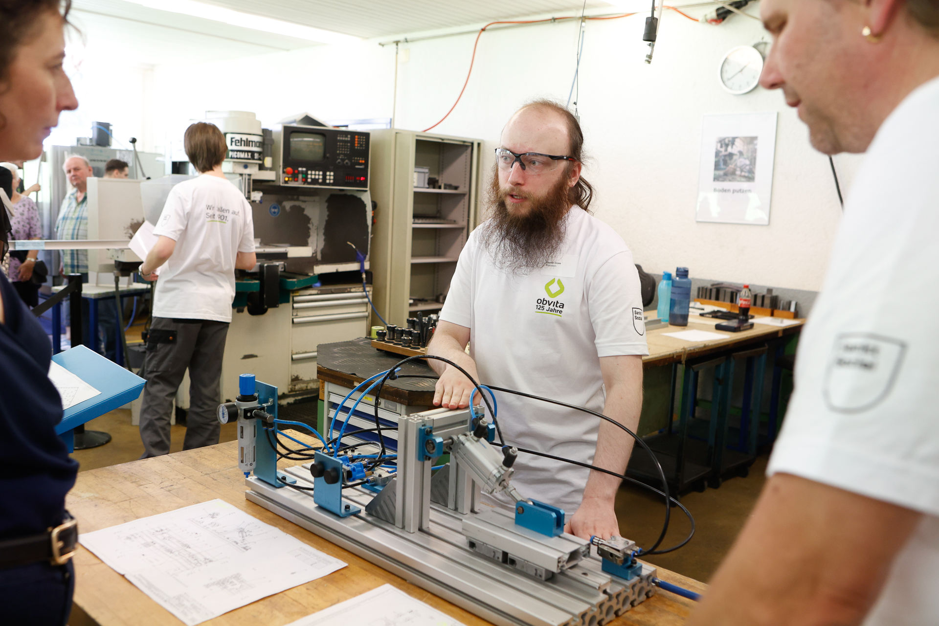 Ein Mann, der eine Schutzbrille und ein weißes T-Shirt trägt, erklärt zwei Personen in einer geschäftigen Werkstatt, in der verschiedene Geräte und Papiere zu sehen sind, die Funktionsweise einer mechanischen Vorrichtung auf einer Werkbank.
