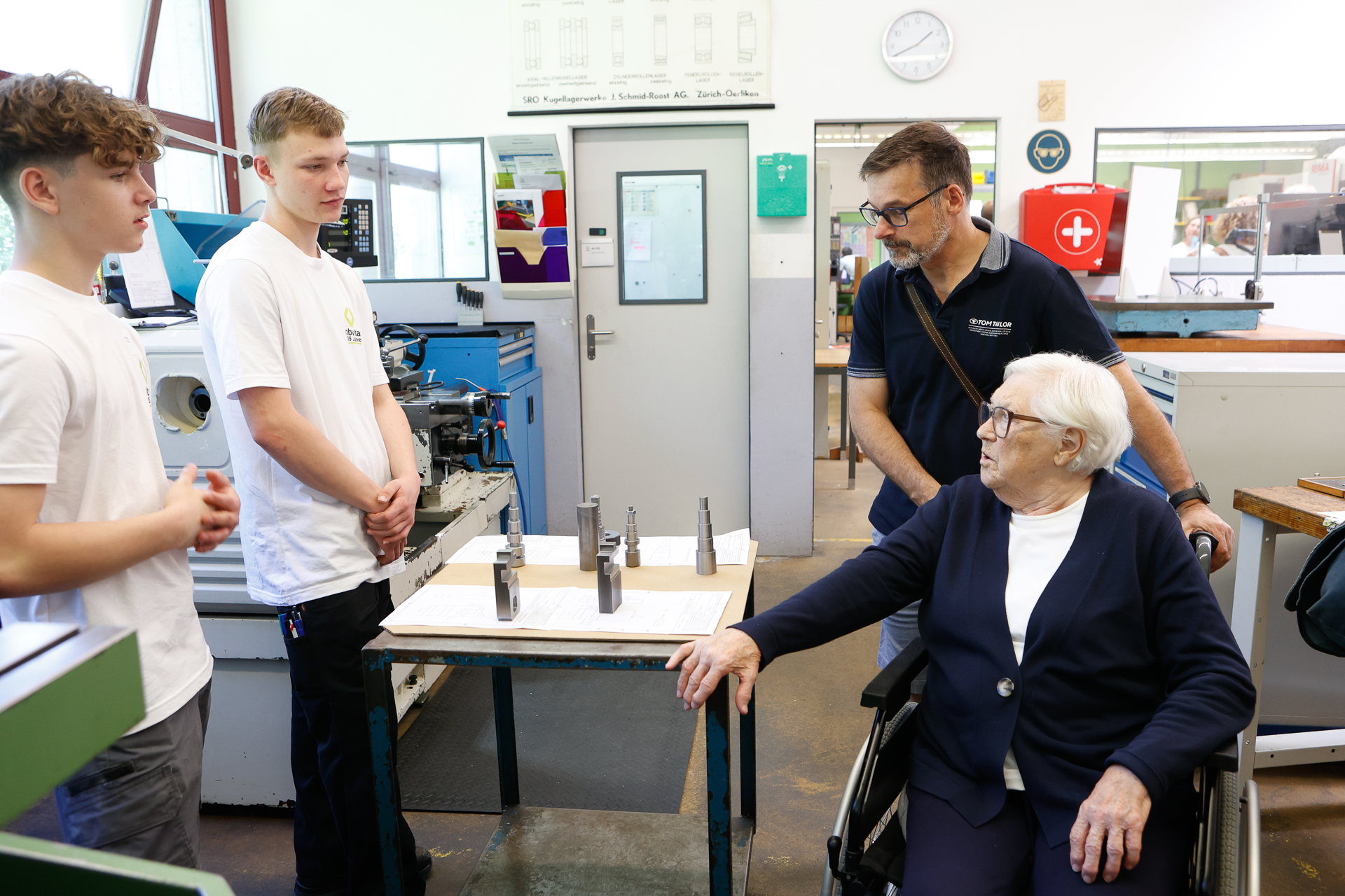 Eine ältere Frau im Rollstuhl unterhält sich mit einem Mann und zwei jungen Männern in einer Werkstatt, die neben einem Tisch mit Metallwerkzeugen stehen. Im Hintergrund sind Maschinen, eine Uhr, Plakate und ein Erste-Hilfe-Kasten zu sehen.