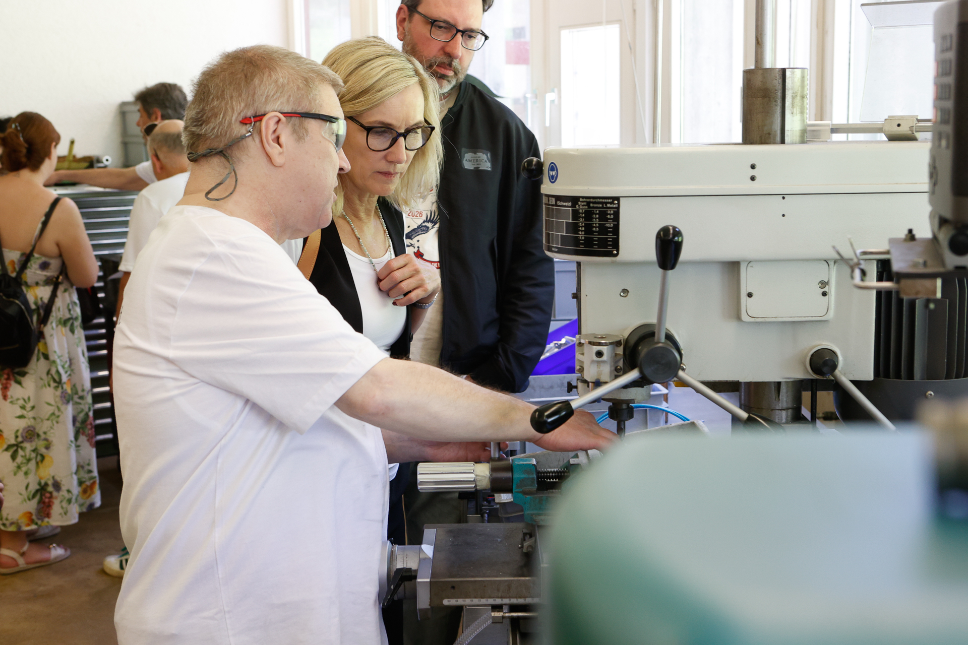 Ein Mann mit Schutzbrille demonstriert zwei aufmerksamen Erwachsenen in einer Werkstattumgebung die Bedienung einer Werkzeugmaschine; im Hintergrund sind weitere Personen und Geräte zu sehen.