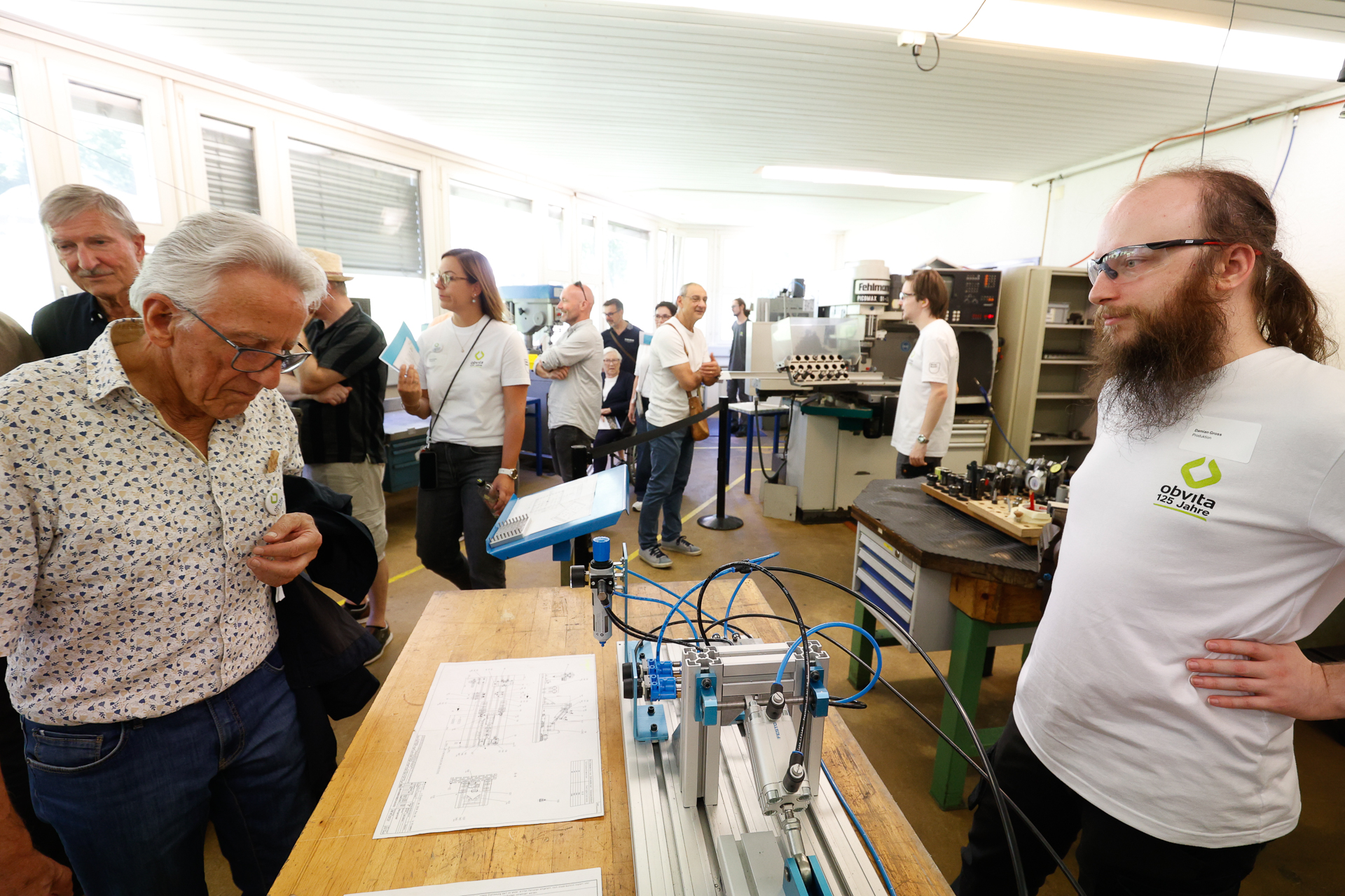 Eine Gruppe von Menschen in einer Werkstatt; ein Mann erklärt einem anderen, älteren Mann die Einstellung einer Maschine, während andere im Hintergrund zuschauen und sich unterhalten. Auf der Werkbank sind technische Zeichnungen und Werkzeuge zu sehen.
