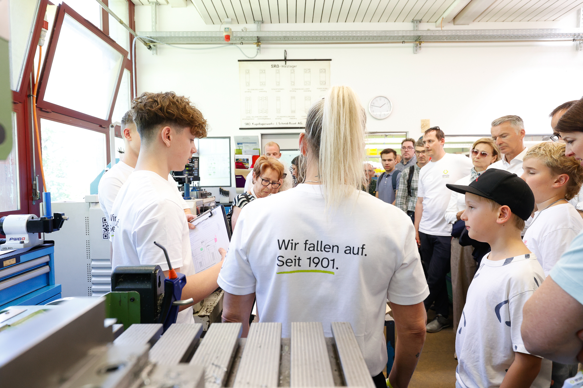 Eine Gruppe von Menschen, die größtenteils weiße Hemden tragen, versammelt sich in einer hellen Werkstatt. Eine Person hält vor der Gruppe einen Vortrag, während die anderen aufmerksam zuhören. Auf dem Hemd steht: „Wir fallen auf. Seit 1901.“ Maschinen und Werkzeuge sind zu sehen.