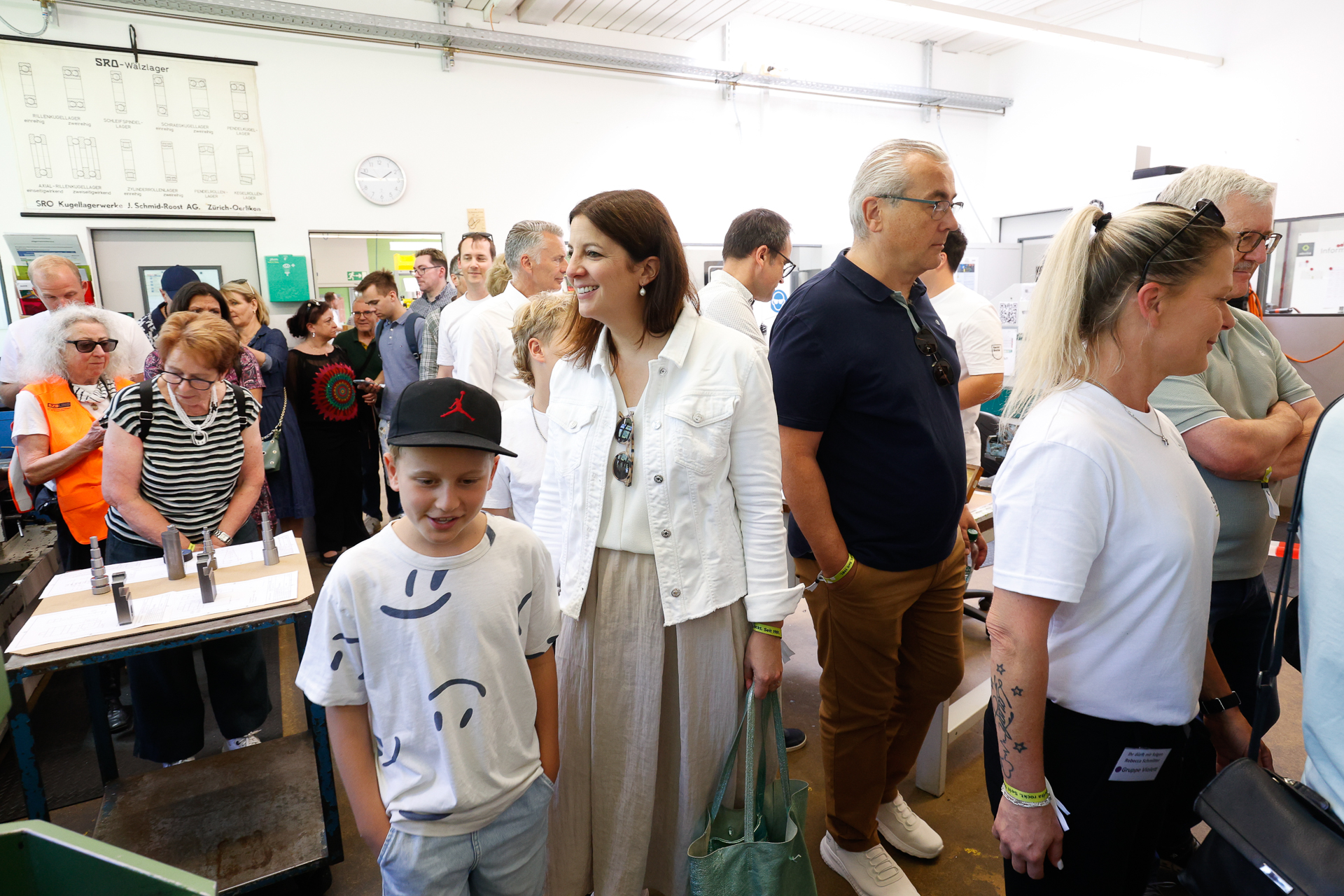 Eine Gruppe von Menschen, darunter Erwachsene und Kinder, steht in einem Raum dicht beieinander, lächelt und unterhält sich. Einige tragen Freizeitkleidung und Armbänder. Der Ort scheint ein Workshop oder eine Veranstaltung mit Tag der offenen Tür zu sein.