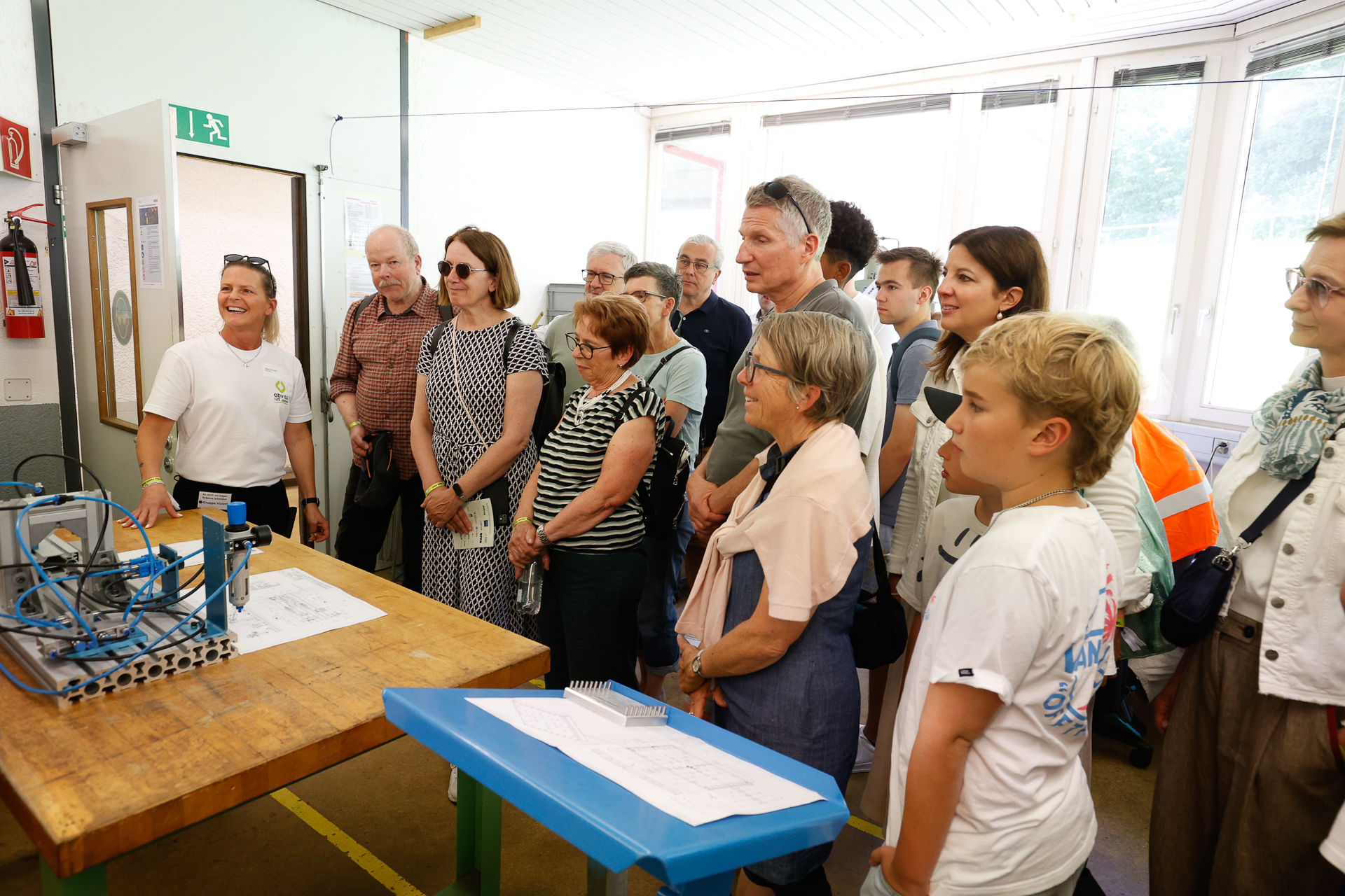 Eine Gruppe von Menschen steht in einer Werkstatt und hört aufmerksam einer Frau zu, die Erklärungen gibt und dabei auf Metallmaschinen und technische Zeichnungen auf einem Tisch zeigt. Die Gruppe wirkt interessiert und konzentriert auf die Vorführung.