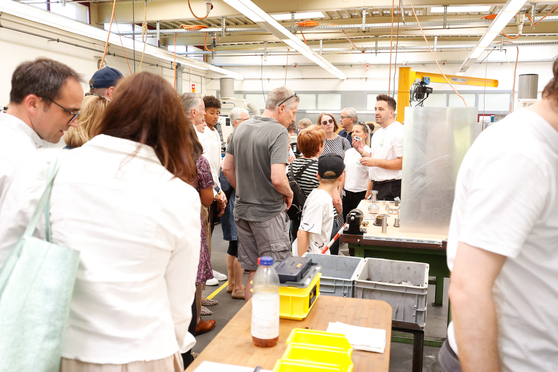 In einer hellen Industriehalle versammelt sich eine Gruppe von Menschen um einen Mann, der eine Maschine erklärt. Erwachsene und Kinder hören aufmerksam zu; auf den Tischen in der Nähe liegen Werkzeuge und Plastikbehälter.