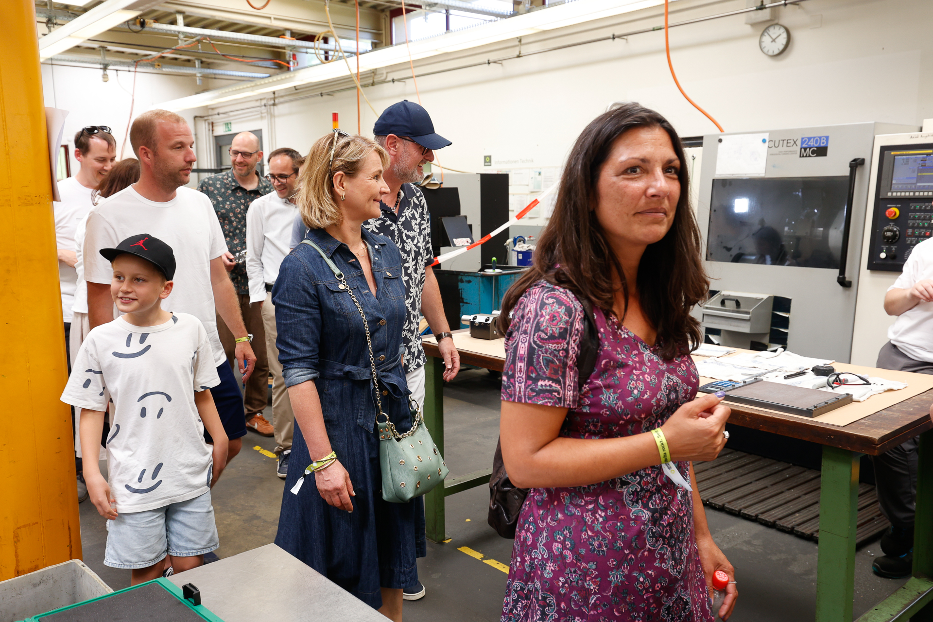 Eine Gruppe von Menschen, darunter Erwachsene und ein Kind, besichtigt eine Produktionsstätte, in der Maschinen und Anlagen zu sehen sind; im Vordergrund steht eine Frau in einem geblümten Kleid, während sich die anderen umsehen und hinter ihr hergehen.