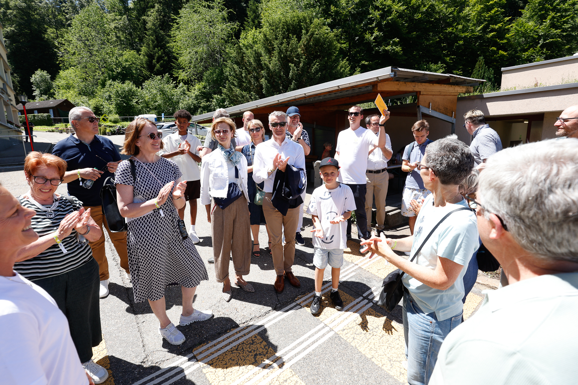 Eine Gruppe von Menschen, darunter Erwachsene und Kinder, steht an einem sonnigen Tag im Freien, lächelt und unterhält sich. Im Hintergrund sind Bäume und kleine Gebäude zu sehen. Einige der Personen halten kleine Gegenstände in der Hand.