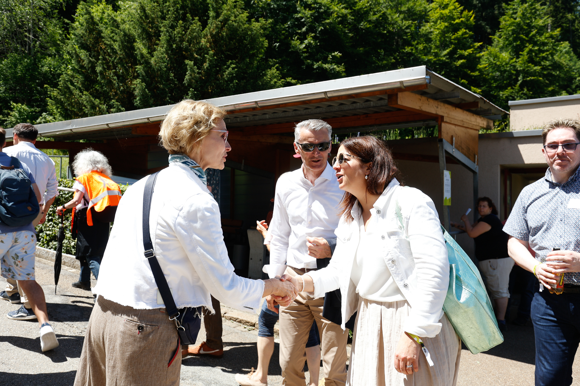 Zwei Frauen geben sich bei einer sonnigen Veranstaltung im Freien die Hand; beide tragen helle Kleidung. In der Nähe stehen und gehen mehrere Menschen, und im Hintergrund sind Bäume zu sehen.
