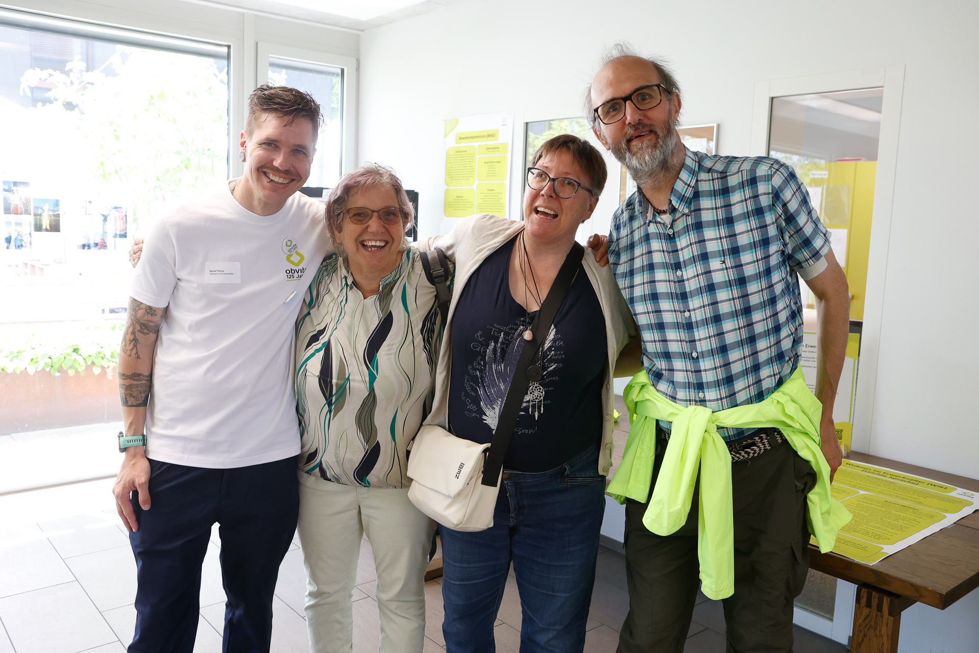 Vier Erwachsene stehen in einem Raum, lächeln und haben ihre Arme umeinander gelegt. Sie wirken glücklich und entspannt. Hinter ihnen sind große Fenster und Informationsplakate an der Wand zu sehen.