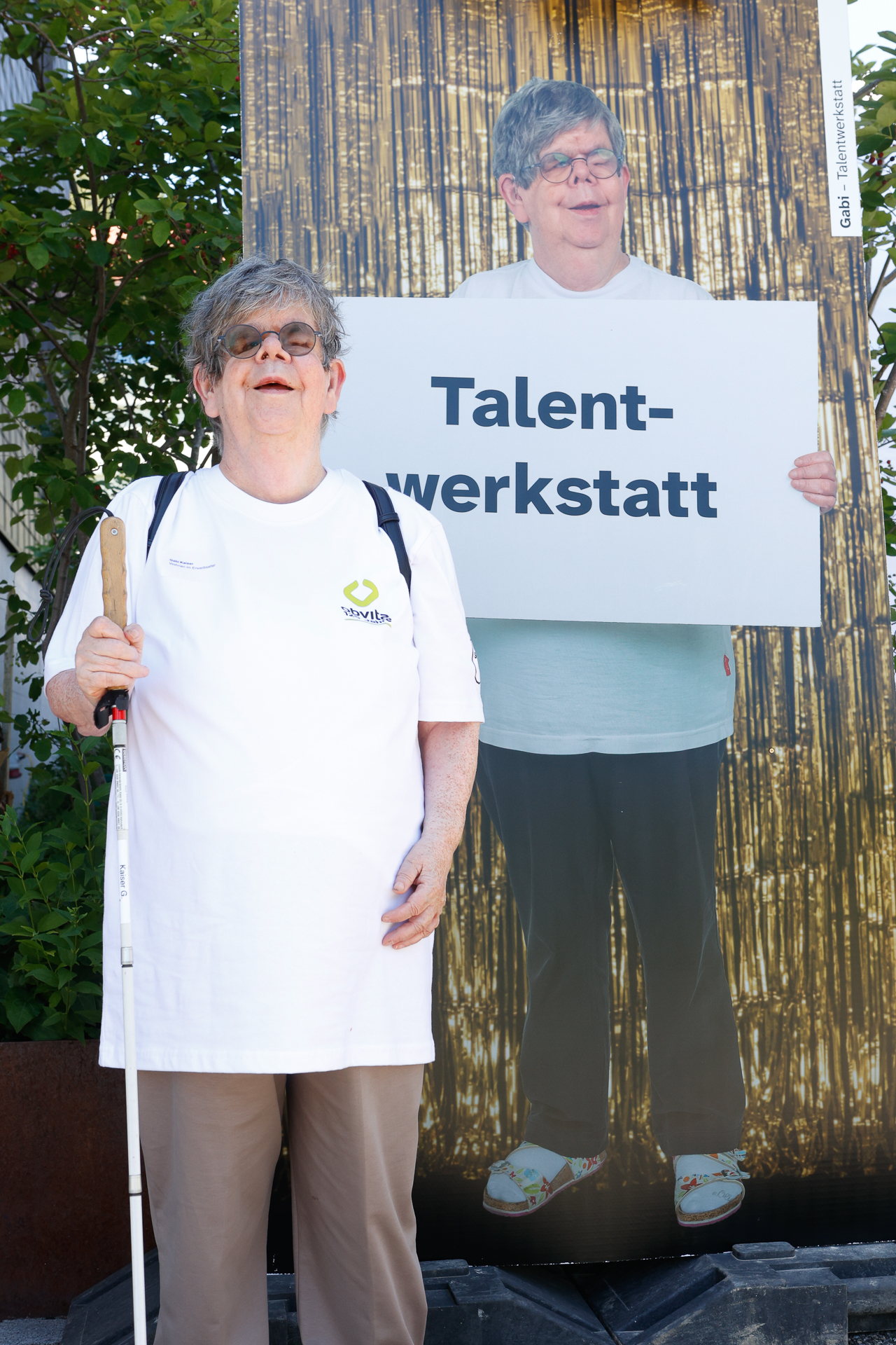 Eine ältere Frau mit Brille und in einem weißen T-Shirt, die einen weißen Blindenstock in der Hand hält, steht neben einem lebensgroßen Plakat, auf dem sie selbst zu sehen ist und ein Schild mit der Aufschrift „Talent-werkstatt“ in der Hand hält. Im Hintergrund ist Grün zu sehen.