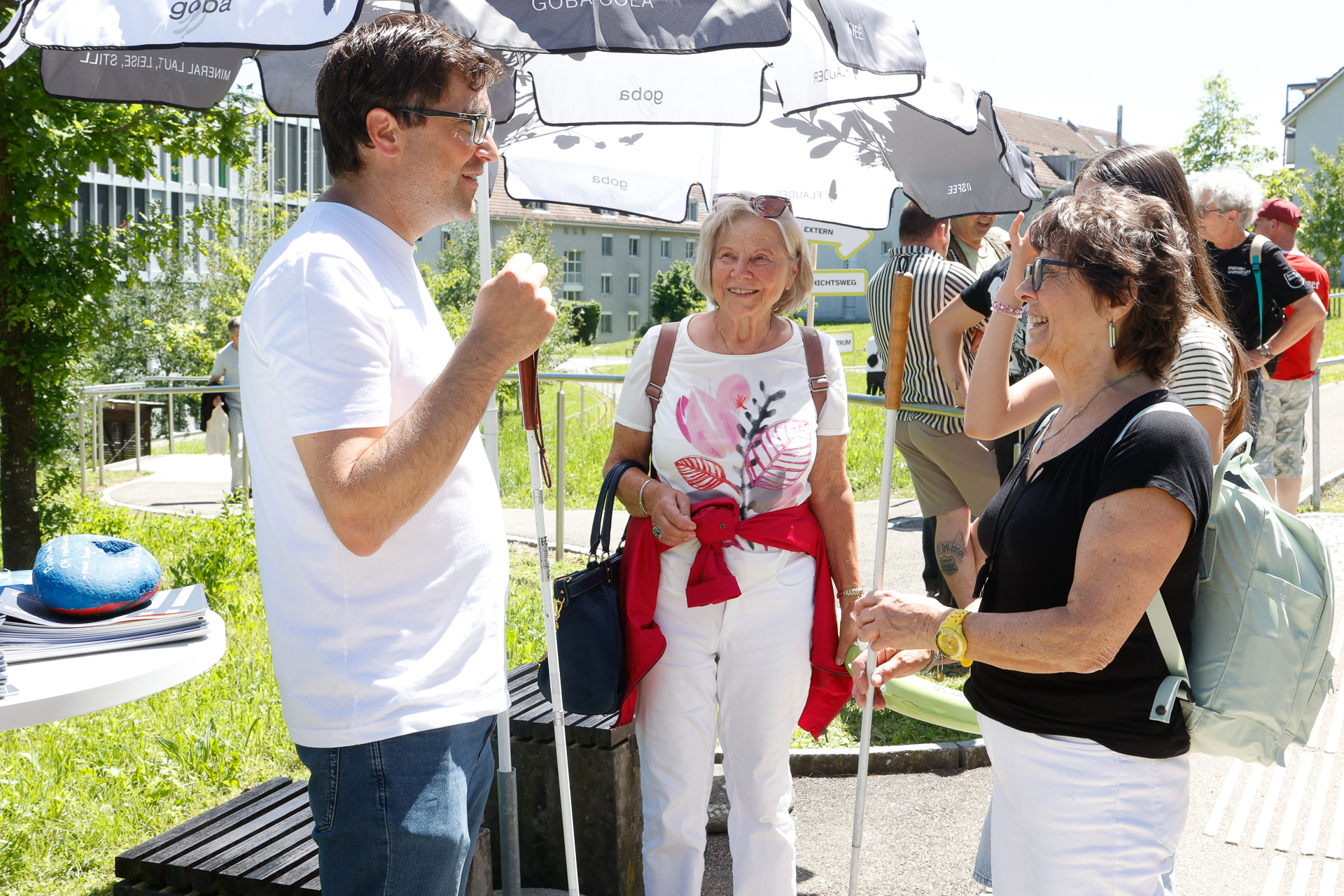 Ein Mann und zwei Frauen stehen an einem sonnigen Tag unter Sonnenschirmen und unterhalten sich. Sie lächeln und sind leger gekleidet; im Hintergrund sind Grünflächen und andere Menschen zu sehen.