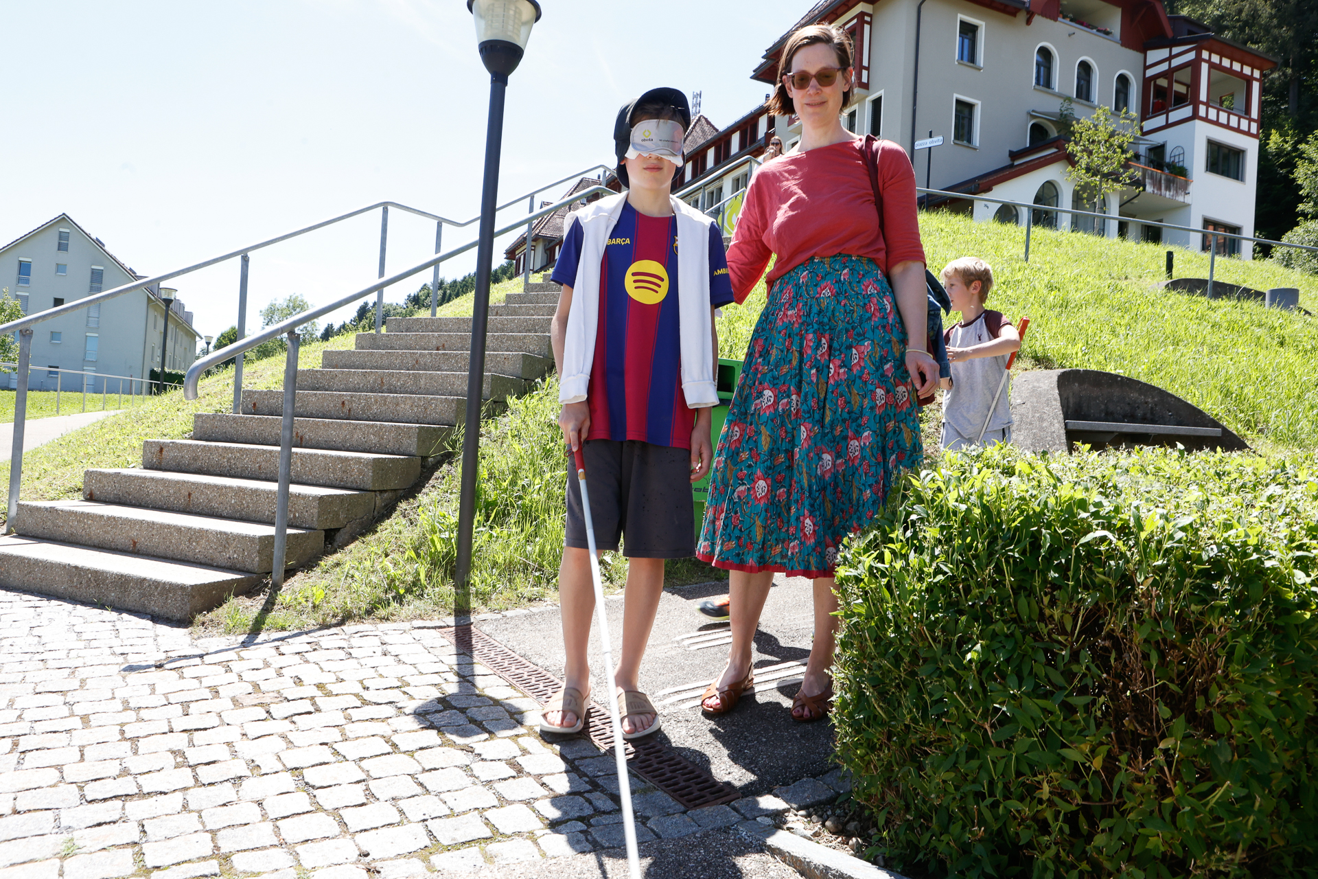 An einem sonnigen Tag steht ein Junge mit einer Augenbinde und einem weißen Blindenstock neben einer Frau in der Nähe einer Treppe und einer Rasenfläche. Ein weiteres Kind steht in der Nähe. Im Hintergrund sind Gebäude zu sehen.