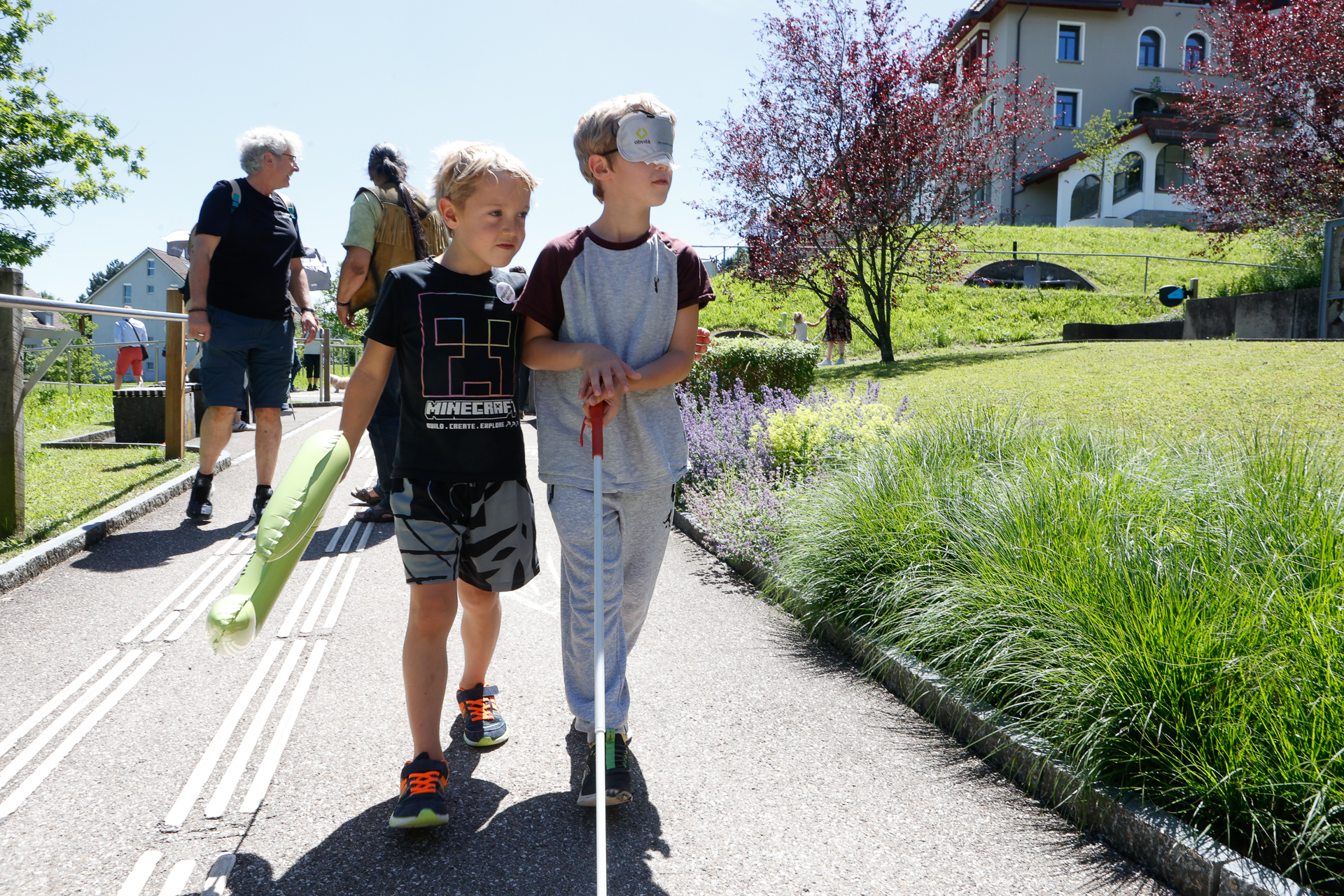 Zwei Jungen gehen auf einem sonnigen Weg; einer hält einen weißen Blindenstock in der Hand und trägt eine Augenbinde, während der andere ihn am Arm führt. Im Hintergrund sind mehrere Menschen zu sehen, die in der Nähe von Wiesen und Häusern spazieren gehen.