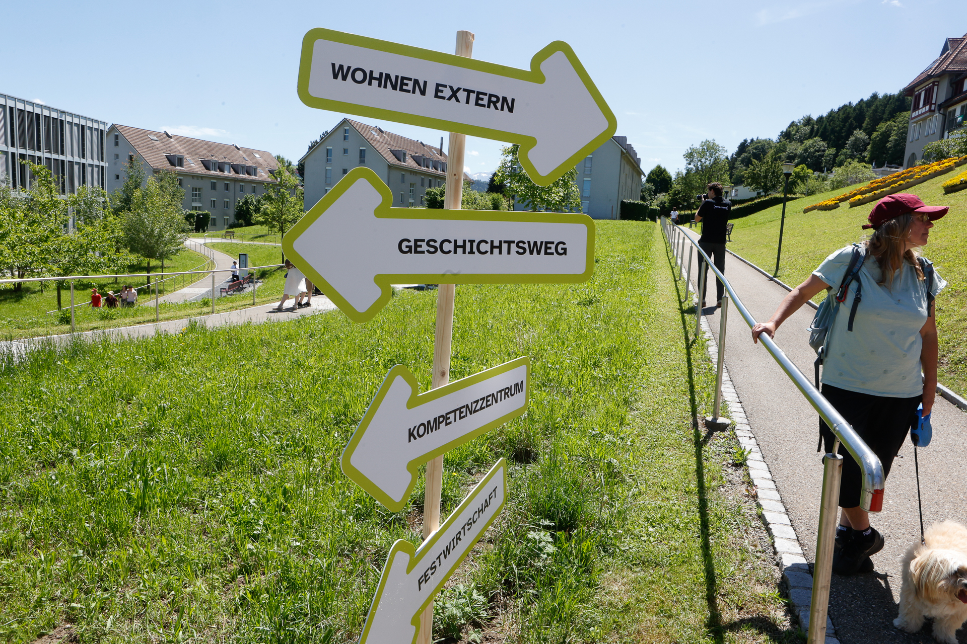An einem sonnigen Tag in einer grünen Wohngegend führt eine Frau einen Hund einen ansteigenden Weg hinauf, neben einem Wegweiser mit vier Pfeilen, auf denen „Wohnen Extern“, „Geschichtsweg“, „Kompetenzzentrum“ und „Festivitätenplatz“ steht.