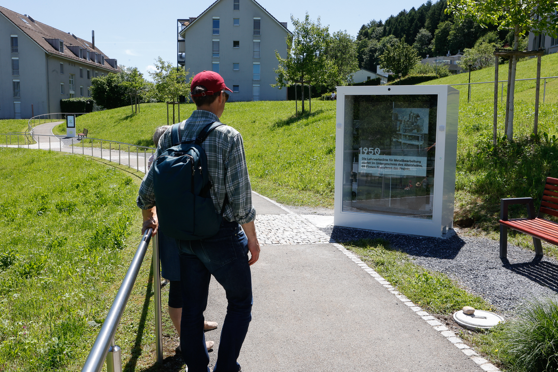 Eine Person mit Rucksack und roter Mütze steht auf einem Weg und betrachtet eine Freiluftausstellung aus der Zeit um 1950. Die Umgebung ist grasbewachsen, in der Nähe befinden sich moderne Gebäude und Bänke, im Hintergrund sind Bäume zu sehen.