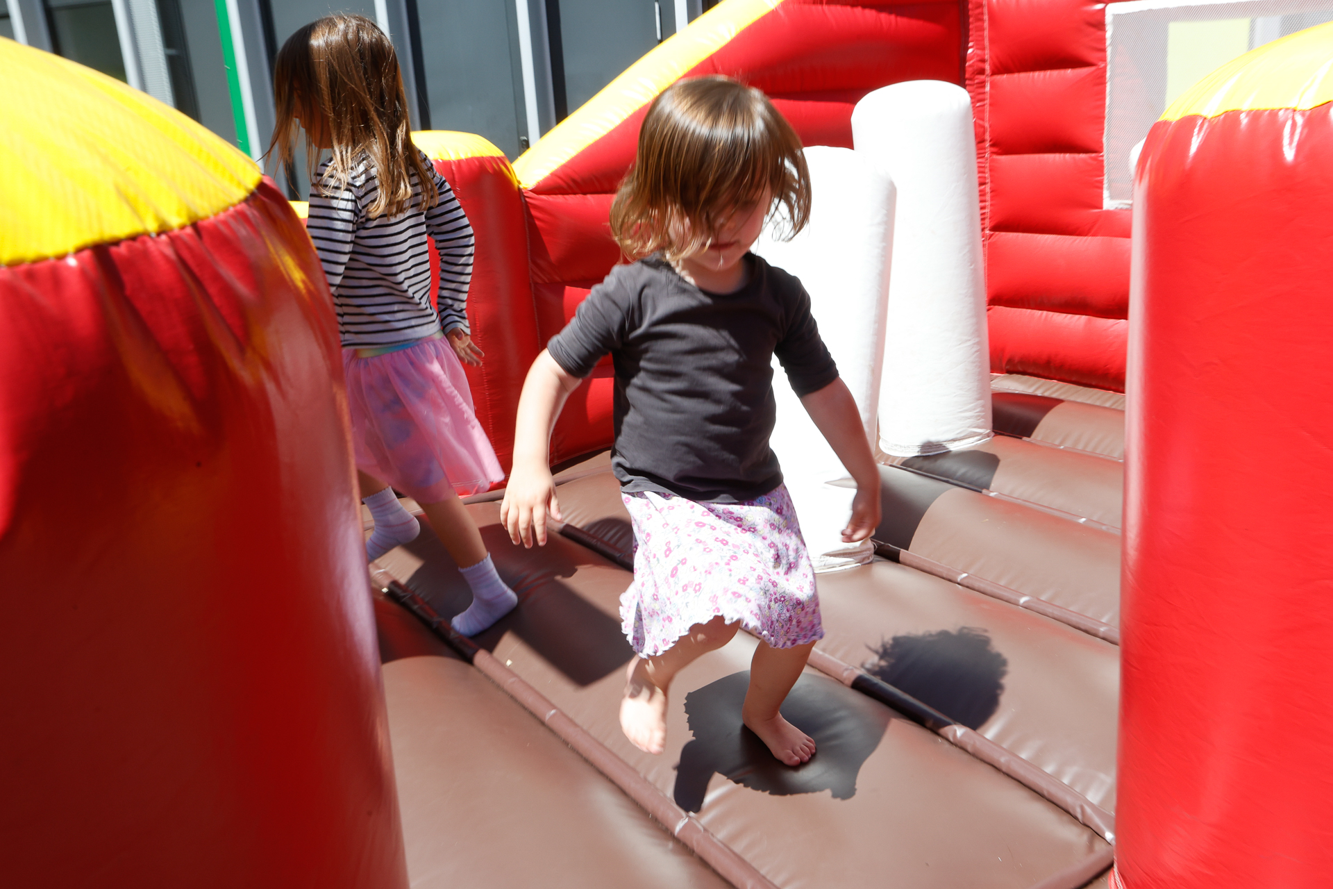 Zwei kleine Kinder spielen barfuß in einer bunten Hüpfburg mit roten Wänden und braunen, gelben und weißen Elementen und genießen den sonnigen Tag.