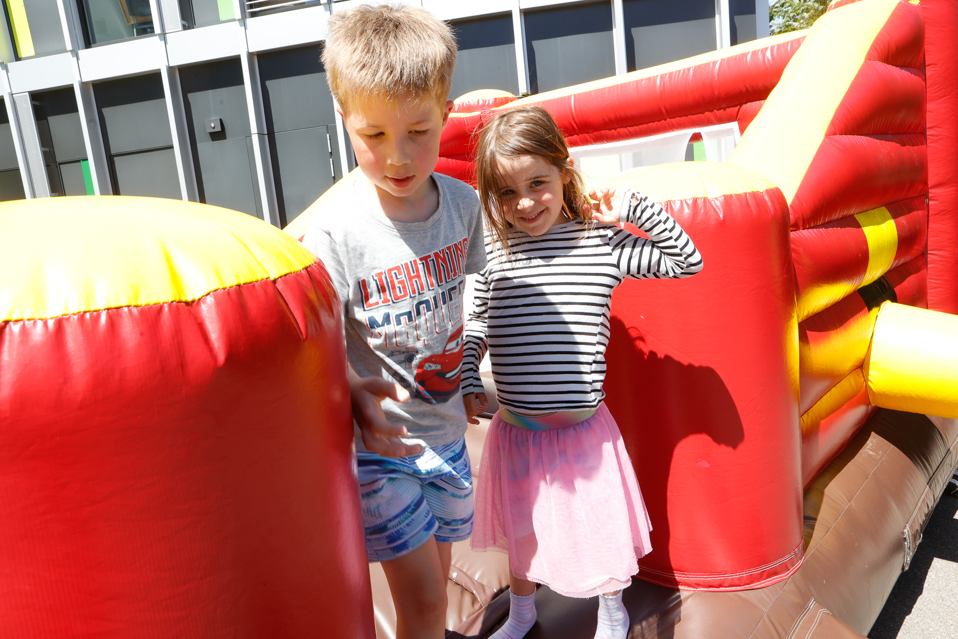 Zwei Kinder stehen draußen auf einem aufblasbaren Hindernisparcours. Der Junge trägt ein graues T-Shirt und blaue Shorts, das Mädchen ein gestreiftes Shirt und einen rosa Rock; beide lächeln und winken. Um sie herum stehen bunte aufblasbare Wände.