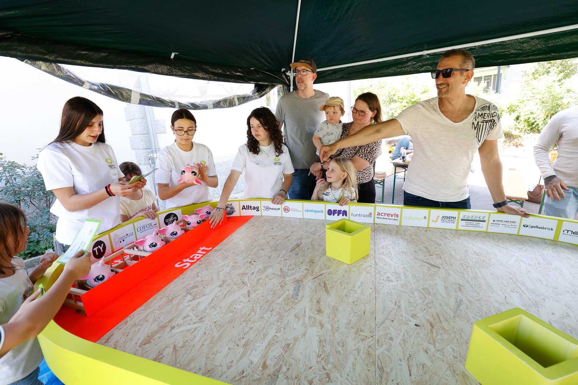 Eine Gruppe aus Erwachsenen und Kindern versammelt sich um einen Tisch mit einer „Start“-Linie und beobachtet kleine Spielzeugschweine, die für ein Rennen aufgestellt wurden. Einige der Anwesenden lächeln, und die Veranstaltung scheint im Freien unter einem Vordach stattzufinden.