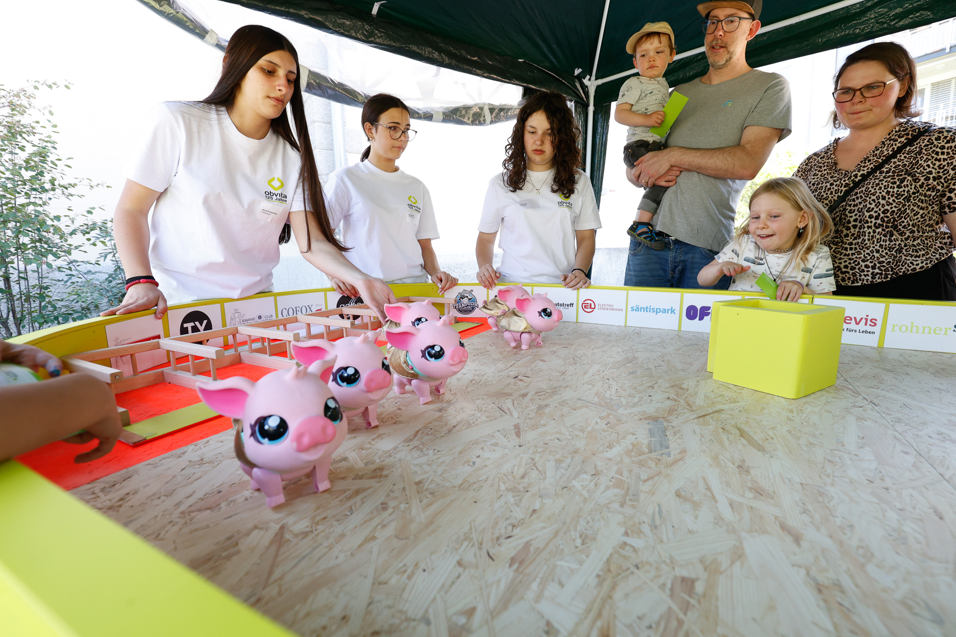 Mehrere Kinder und Erwachsene versammeln sich um einen Tisch mit rosa Spielzeug-Schwein-Robotern in einer kleinen, eingezäunten Arena, während andere unter einem Zelt im Freien zuschauen und helfen.