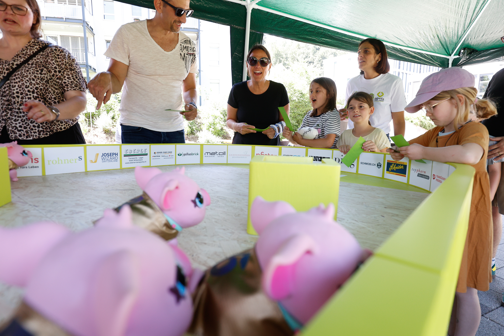 Kinder und Erwachsene lächeln und schauen zu, wie mehrere rosa Spielzeugschweine bei einer Veranstaltung im Freien in einem kleinen, umzäunten Ring unter einem Vordach um die Wette laufen. Die Gruppe wirkt begeistert und ist ganz in die Aktivität vertieft.