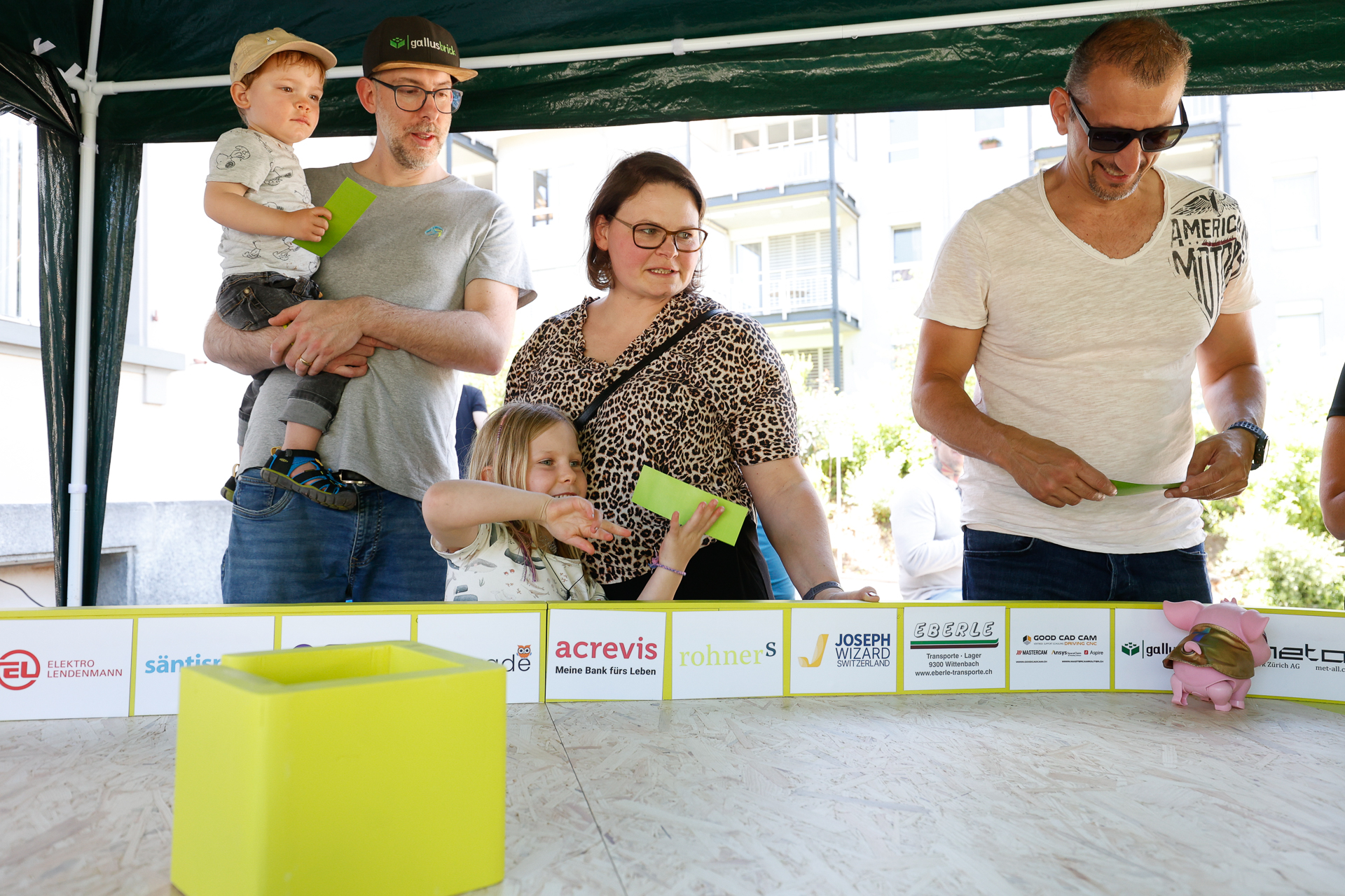 Eine Gruppe aus Erwachsenen und Kindern steht unter einem Zelt, hält grüne Karten in den Händen und blickt auf einen Spieltisch mit gelben Gegenständen. Die Kulisse lässt auf eine ungezwungene Veranstaltung im Freien oder einen Jahrmarkt schließen.