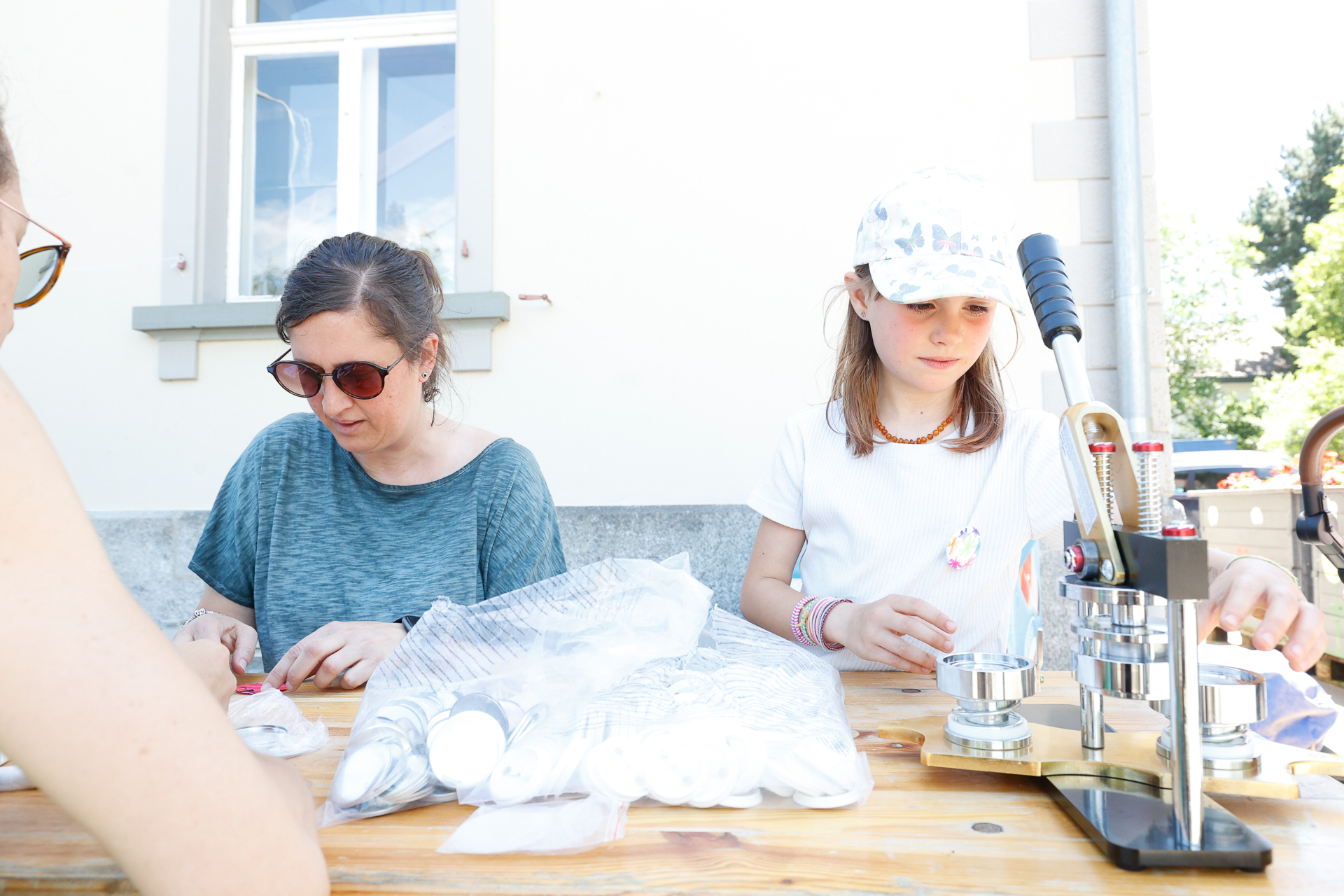 Zwei Personen sitzen draußen an einem Holztisch und stellen mit einer Knopfpresse Knöpfe her. Eine Person trägt eine Sonnenbrille und ein blaues Hemd, die andere eine weiße Mütze und ein weißes Hemd. Auf dem Tisch liegen mehrere Knopfrohlinge und Materialien ausgebreitet.