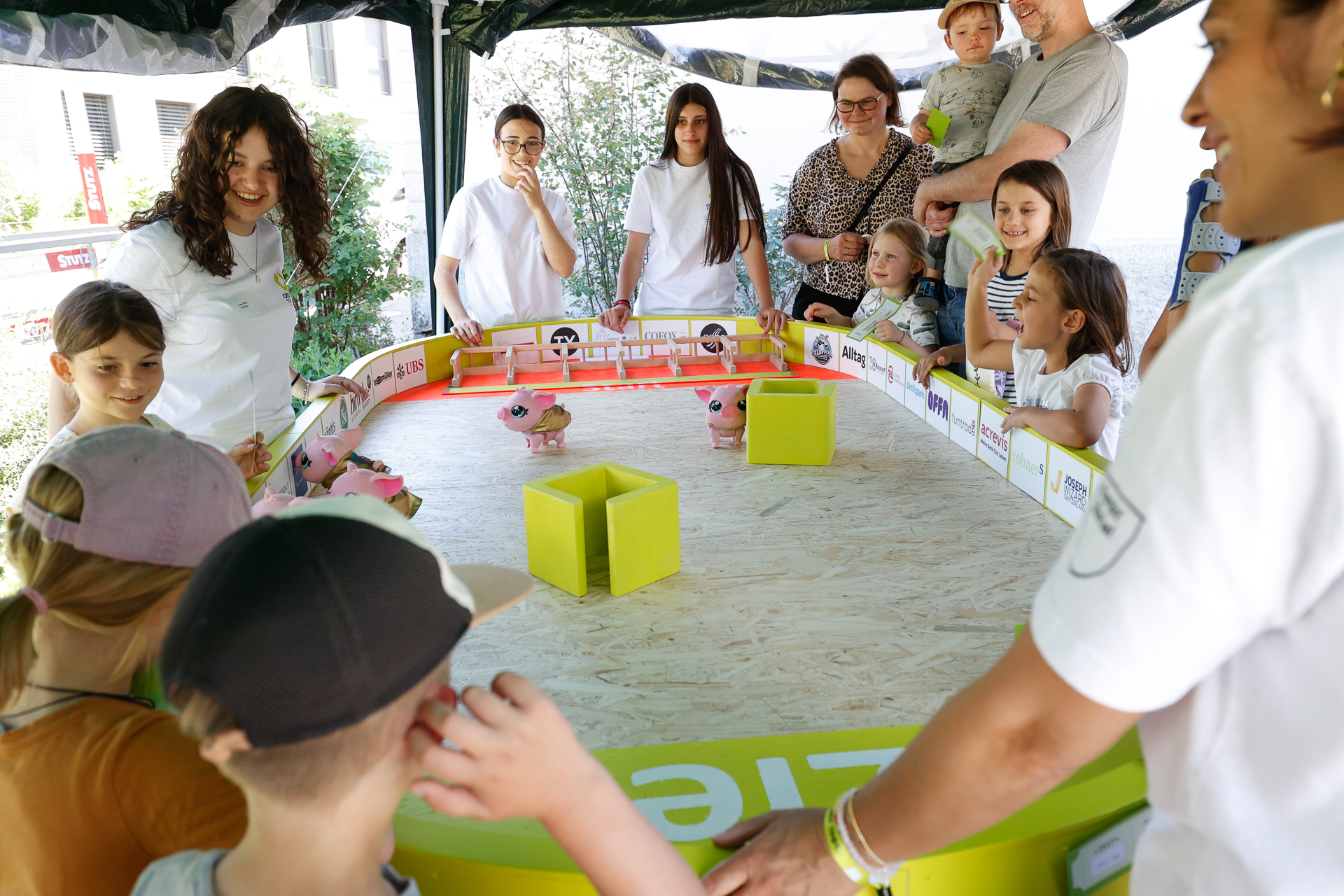 Eine Gruppe von Kindern und Erwachsenen versammelt sich um eine Arena auf einem Tisch und beobachtet, wie kleine rosa Robotertiere um Hindernisse herumlaufen. Die Szene spielt sich im Freien unter einem Vordach ab, und alle wirken begeistert und glücklich.