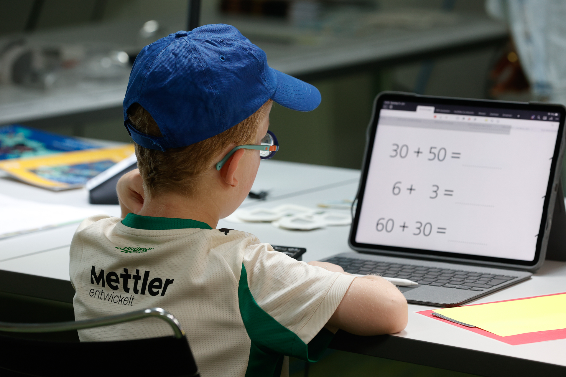 Ein kleiner Junge mit einer blauen Mütze und einer Brille sitzt an einem Schreibtisch und schaut auf ein Tablet, auf dem Mathematikaufgaben angezeigt werden. Auf dem Schreibtisch liegen Schulsachen, und auf dem Bildschirm sind mathematische Gleichungen zu sehen.