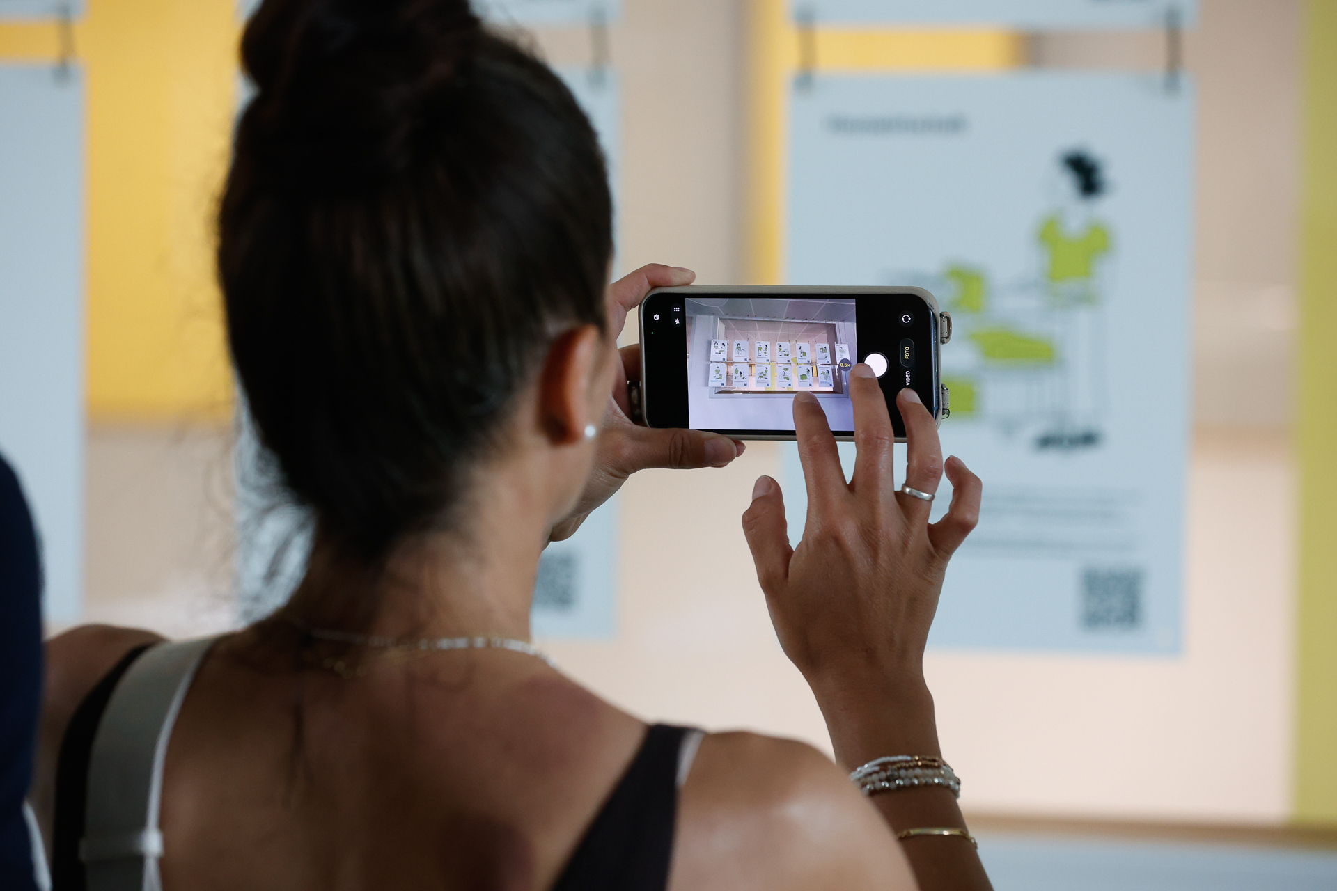 Eine Frau mit dunklem Haar, das zu einem Dutt zusammengebunden ist, fotografiert mit ihrem Smartphone Plakate, die in einem Ausstellungsraum ausgestellt sind. Die Plakate zeigen Illustrationen und Text, und auf einem davon ist ein QR-Code zu sehen.