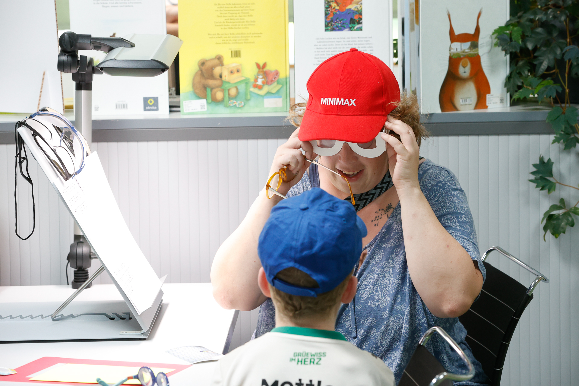 Eine lächelnde Frau mit einer roten „MINIMAX“-Kappe und einer großen Spielzeugbrille sitzt an einem Tisch einem Kind mit einer blauen Kappe gegenüber; im Hintergrund sind bunte Bücher und Kunstwerke zu sehen.