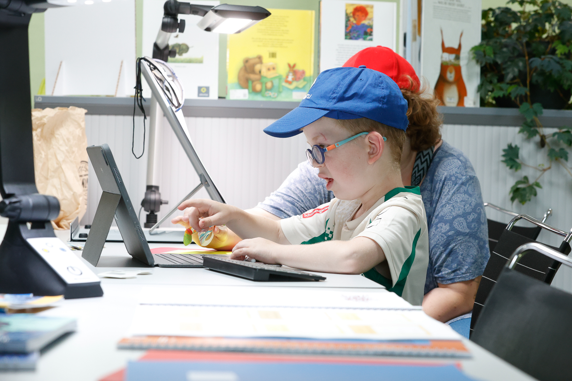 Ein kleines Kind mit Brille und blauer Mütze bedient mit Hilfe eines Erwachsenen ein Tablet in einem Klassenzimmer oder einer Bibliothek. Auf dem Tisch liegen Bücher und Lernmaterialien.