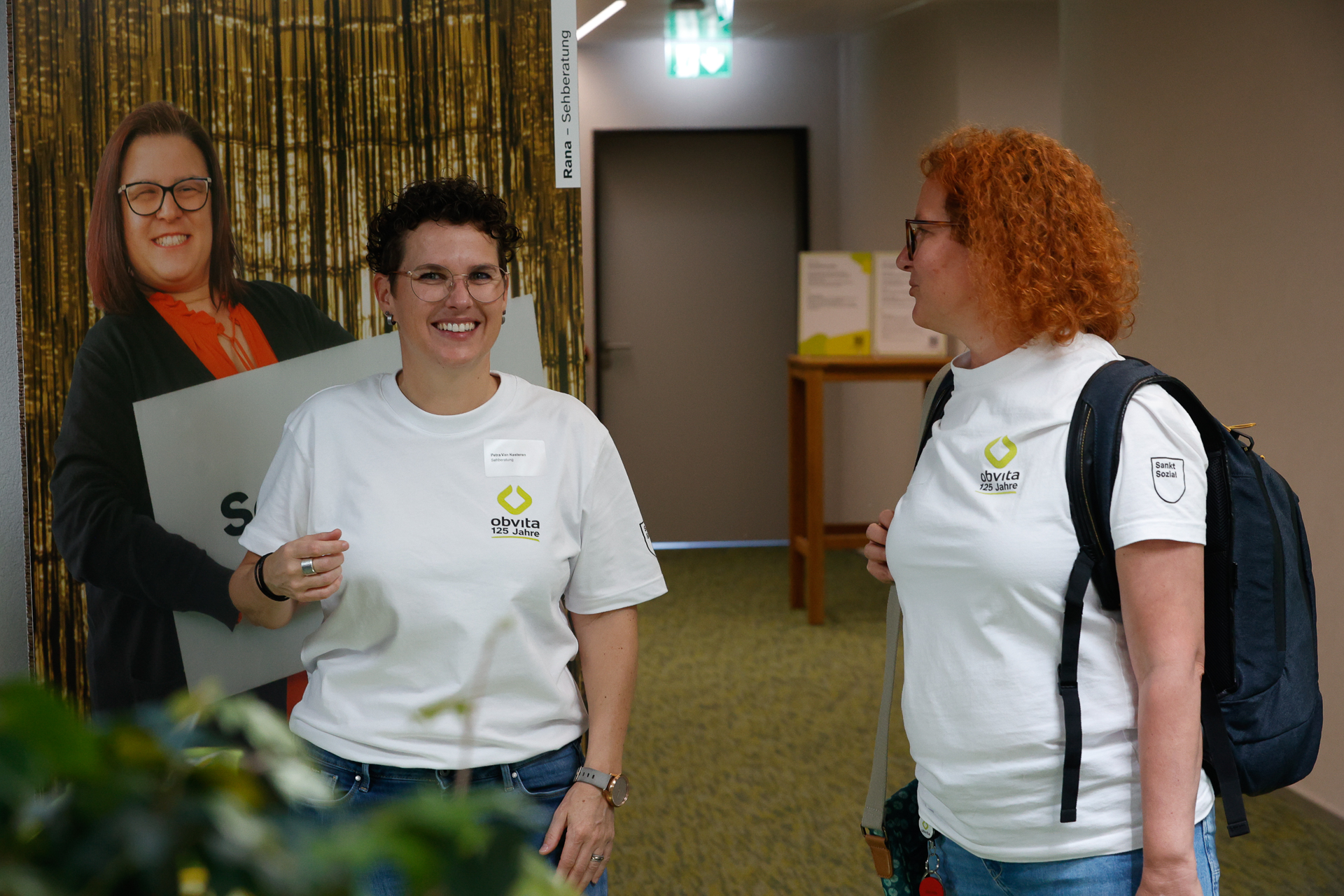 Zwei Frauen in weißen Olyvita-T-Shirts stehen in einem Raum, lächeln und unterhalten sich. Eine Frau steht neben einer Pappfigur einer anderen Frau, die ein Schild in der Hand hält. Im Hintergrund sind eine Tür und Pflanzen zu sehen.