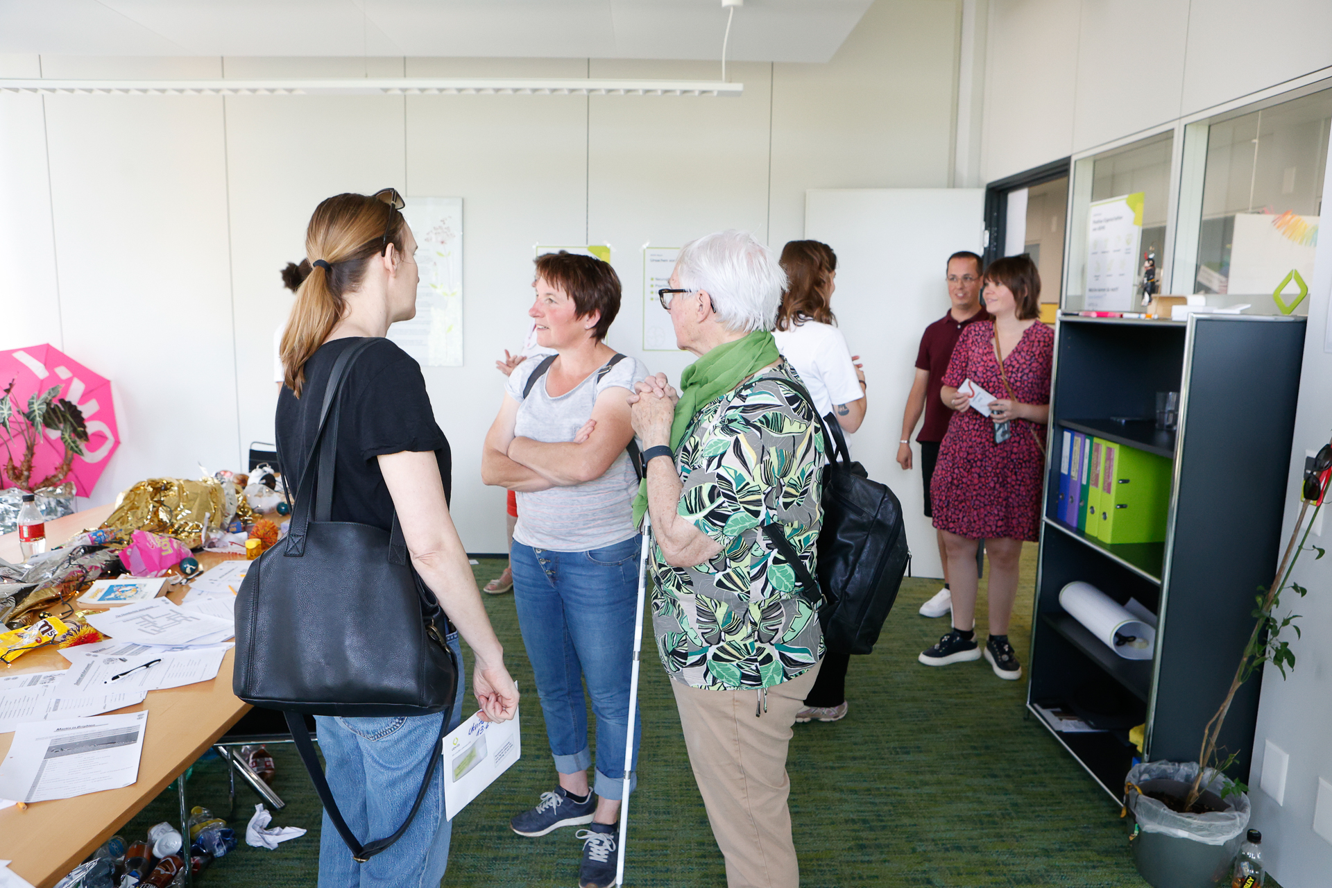 Eine Gruppe von Menschen steht in einem Büro mit einem überfüllten Schreibtisch, Regalen und Papieren und unterhält sich. Im Vordergrund unterhalten sich drei Frauen, während sich im Hintergrund andere unterhalten. Die Atmosphäre wirkt ungezwungen und freundlich.