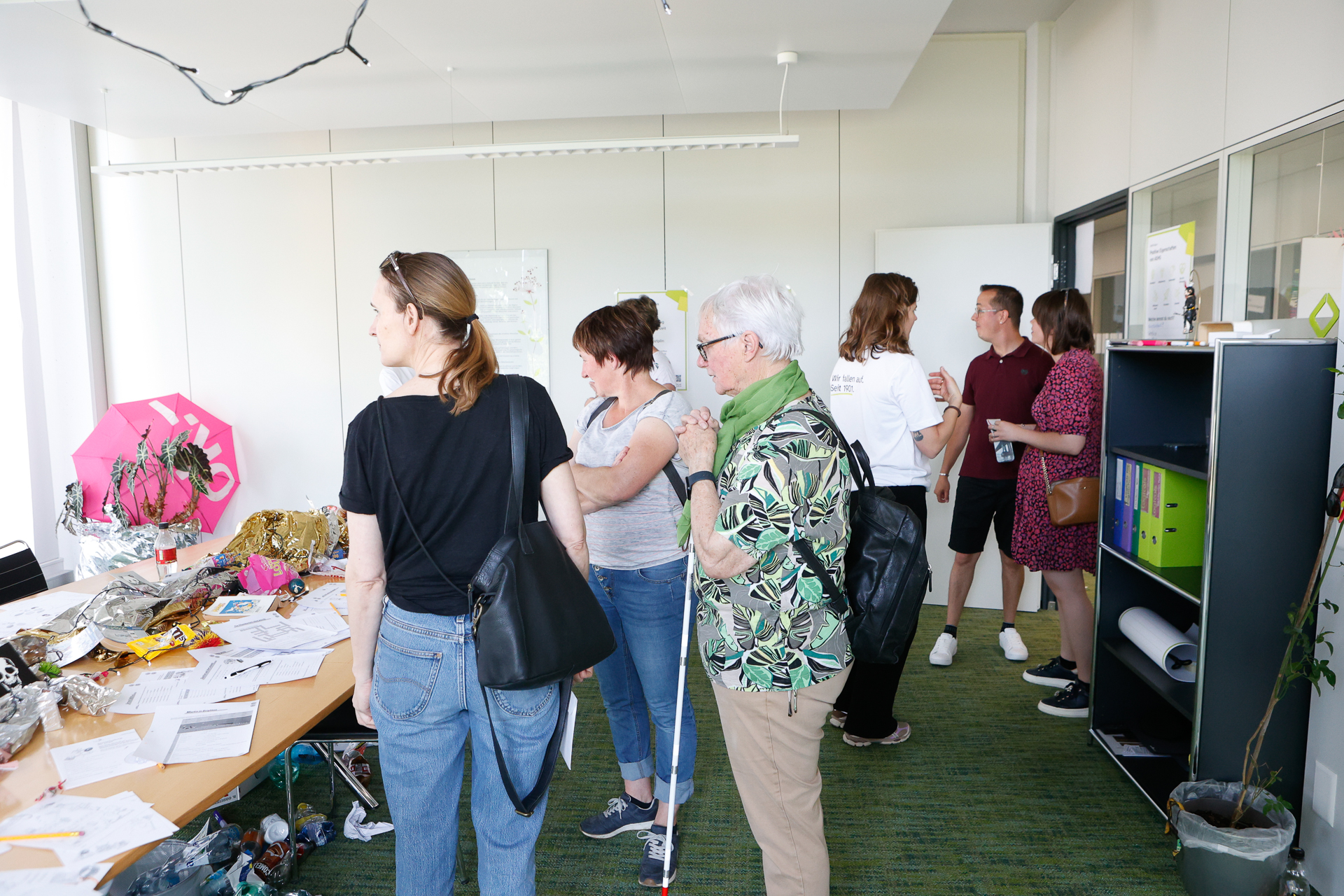 Eine Gruppe von Menschen steht in einem hellen Büroraum und unterhält sich. Auf einem Tisch liegen Papiere und Gegenstände verstreut, und im Hintergrund sind Regale mit Ordnern zu sehen. Die Atmosphäre wirkt locker und ungezwungen.